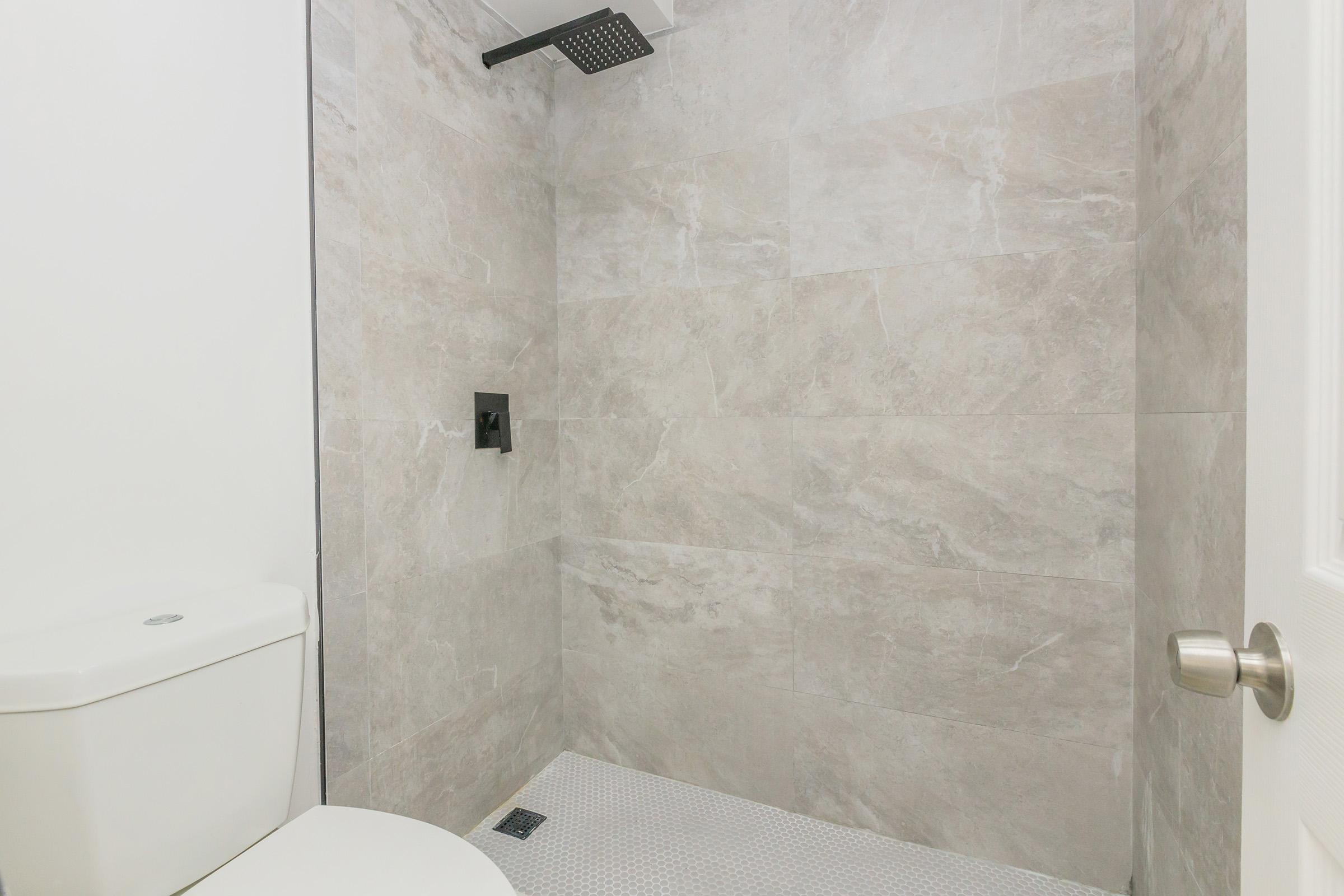 A clean, modern bathroom featuring a shower with a rainfall showerhead, light-colored tiled walls, and a white toilet. The shower area is minimalist and spacious, with neutral tones and a simple, functional design.