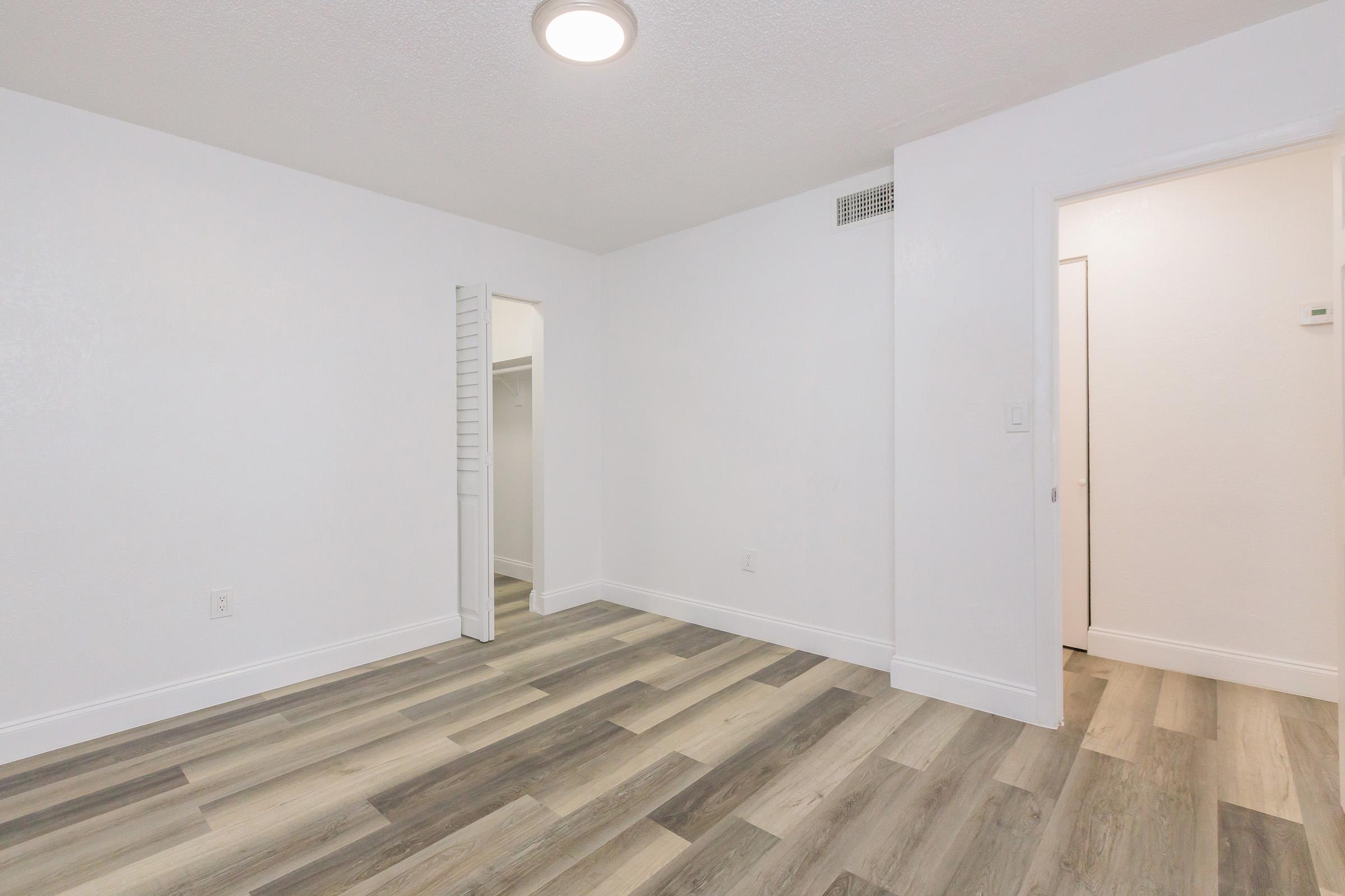 A clean, empty room with white walls and a circular ceiling light. The flooring is a mix of light and dark wood textures. There is a small closet with sliding doors on the left and a doorway leading to another room on the right, creating a bright and spacious atmosphere.