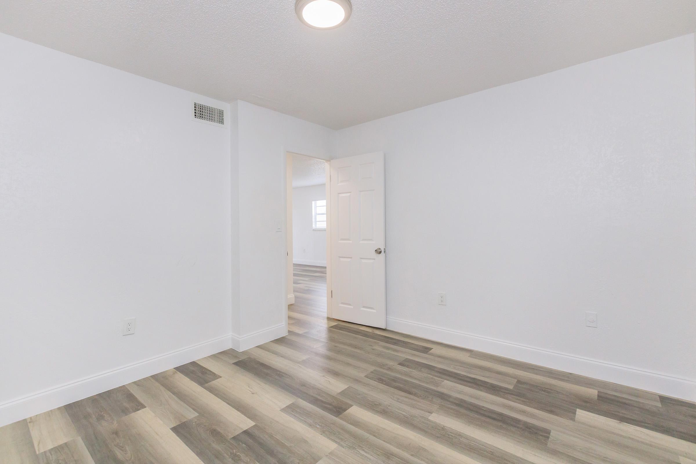 A clean, empty room with light-colored walls and a wood-like floor. A partially open door leads to another room, and a ceiling light fixture provides illumination. The space is well-lit and has a minimalist design, making it suitable for various purposes such as a bedroom or office.
