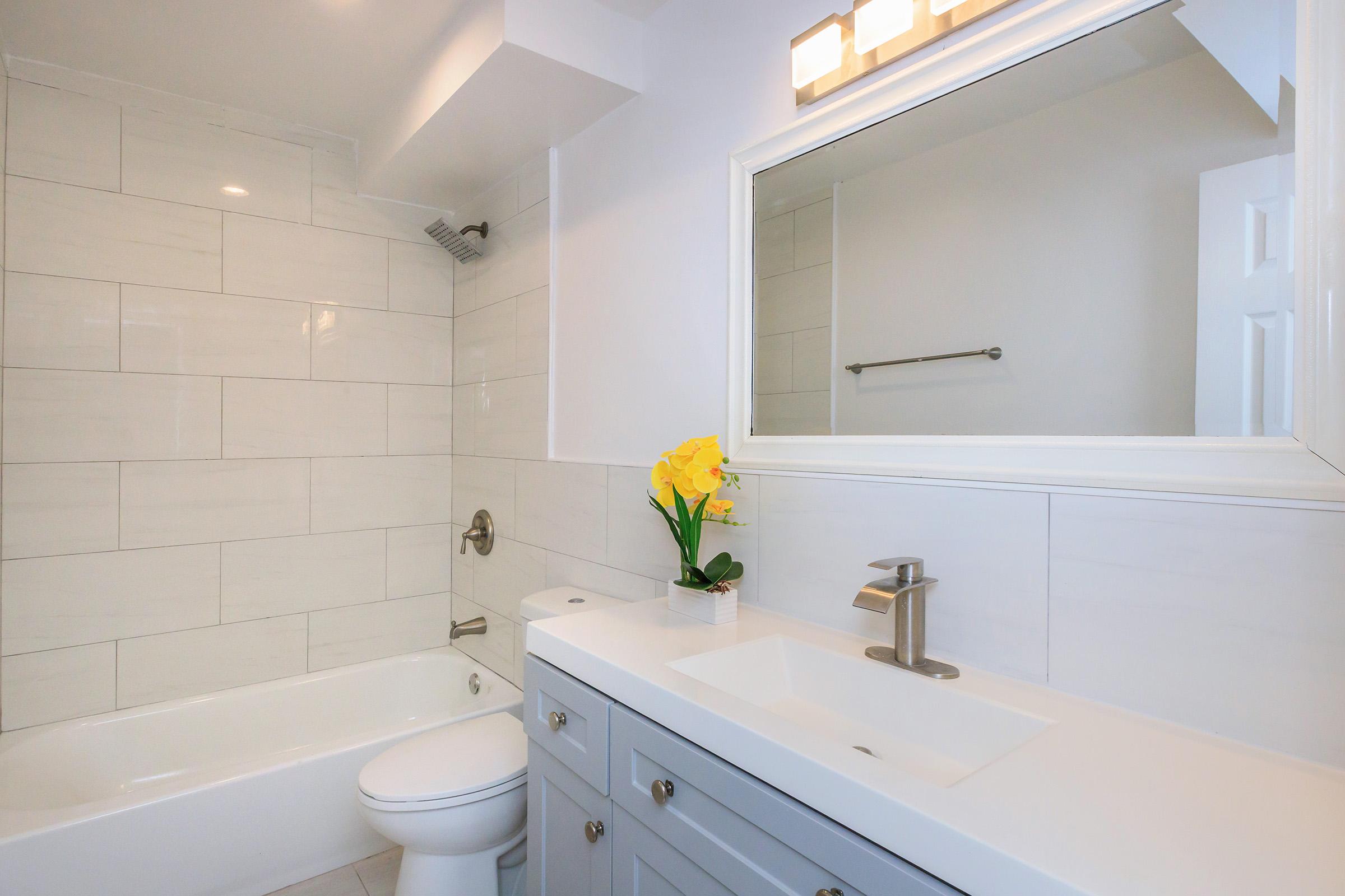 A modern bathroom featuring a white tile shower and bath, a sleek vanity with a sink and faucet, a large mirror above the vanity, and a small vase with yellow flowers on the counter. Soft lighting highlights the neutral color scheme and clean design, creating a bright and inviting atmosphere.