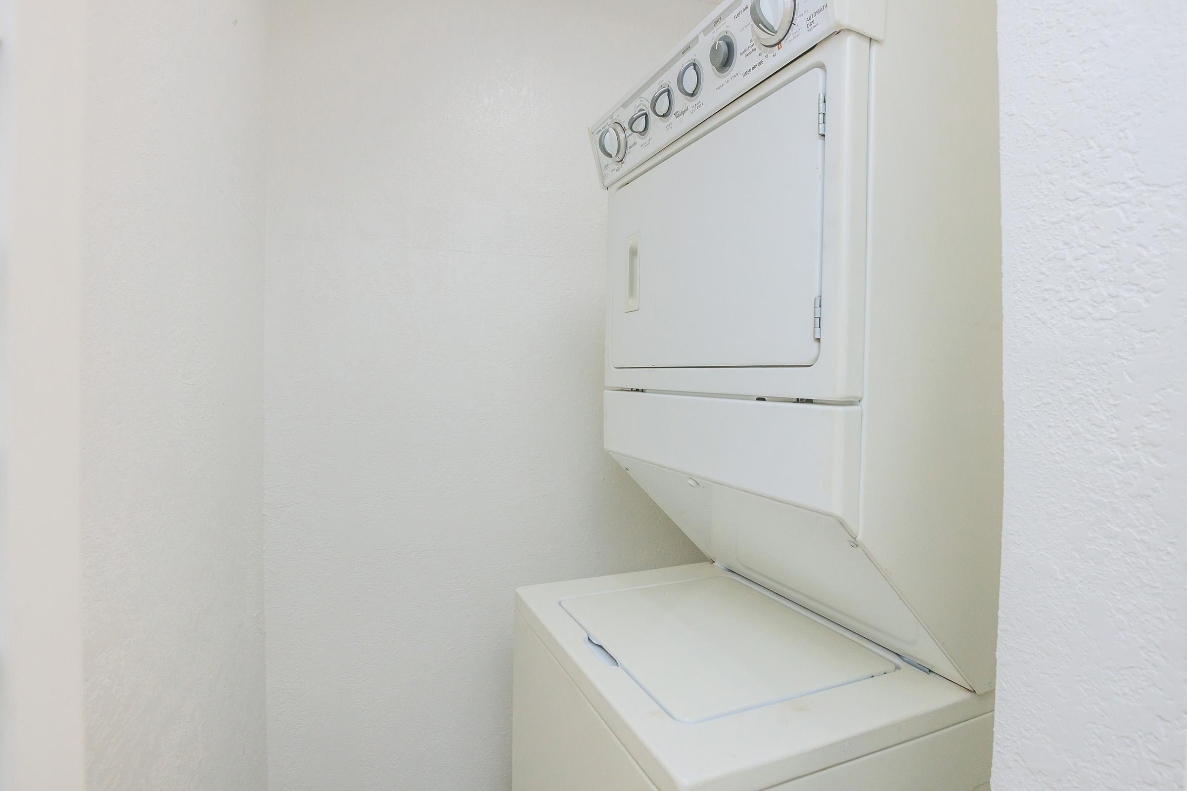 A compact stackable washer and dryer located in a small, white-walled laundry area. The appliances are white and positioned one on top of the other, with controls visible on the front of the dryer. The space appears clean and minimalistic, suitable for laundry tasks.