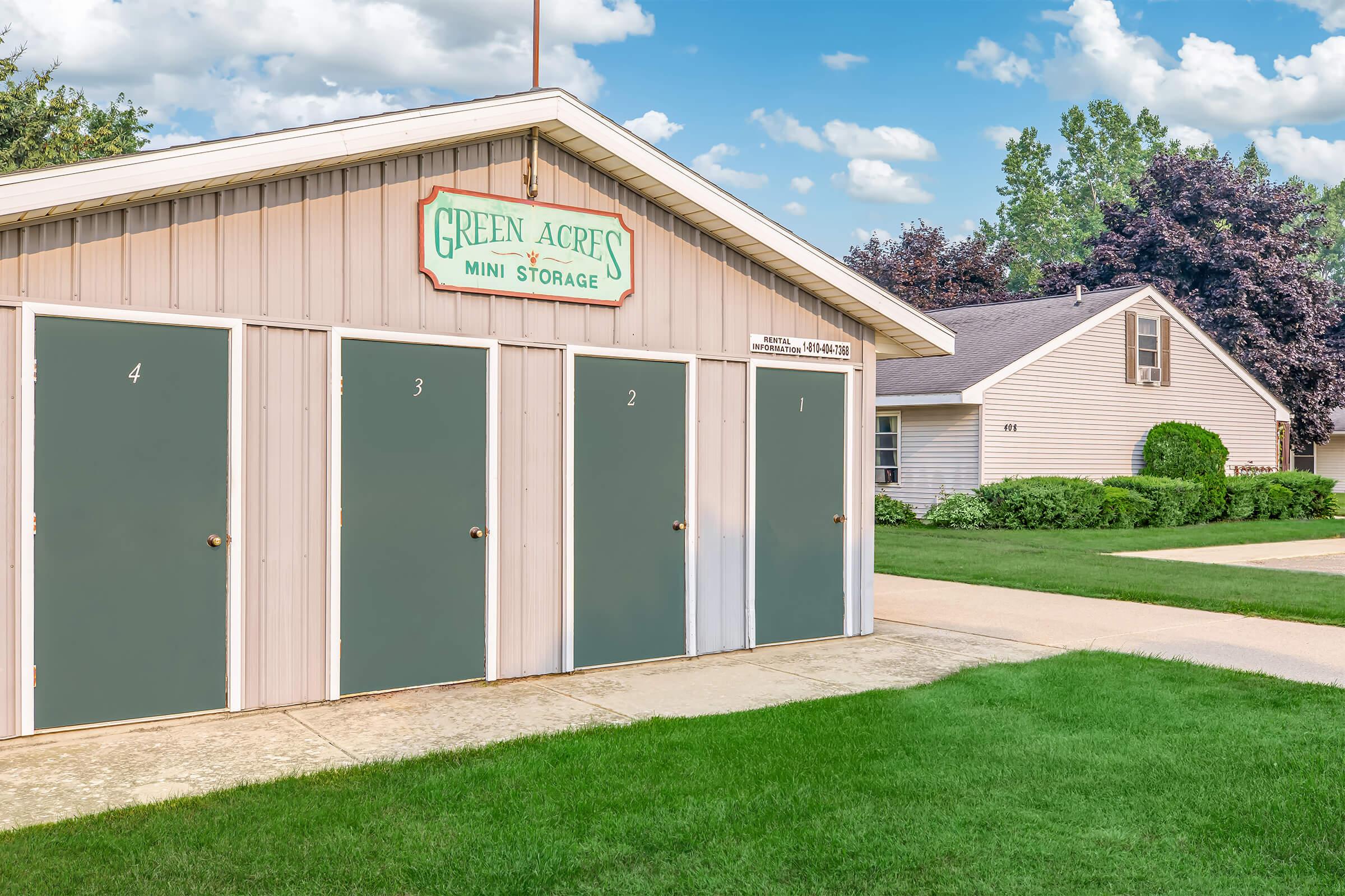 A small storage facility with four green doors labeled 1, 2, 3, and 4. The building has a sign that reads "Green Acres Mini Storage." In the background, there is a residential house and well-maintained green lawns under a partly cloudy sky.