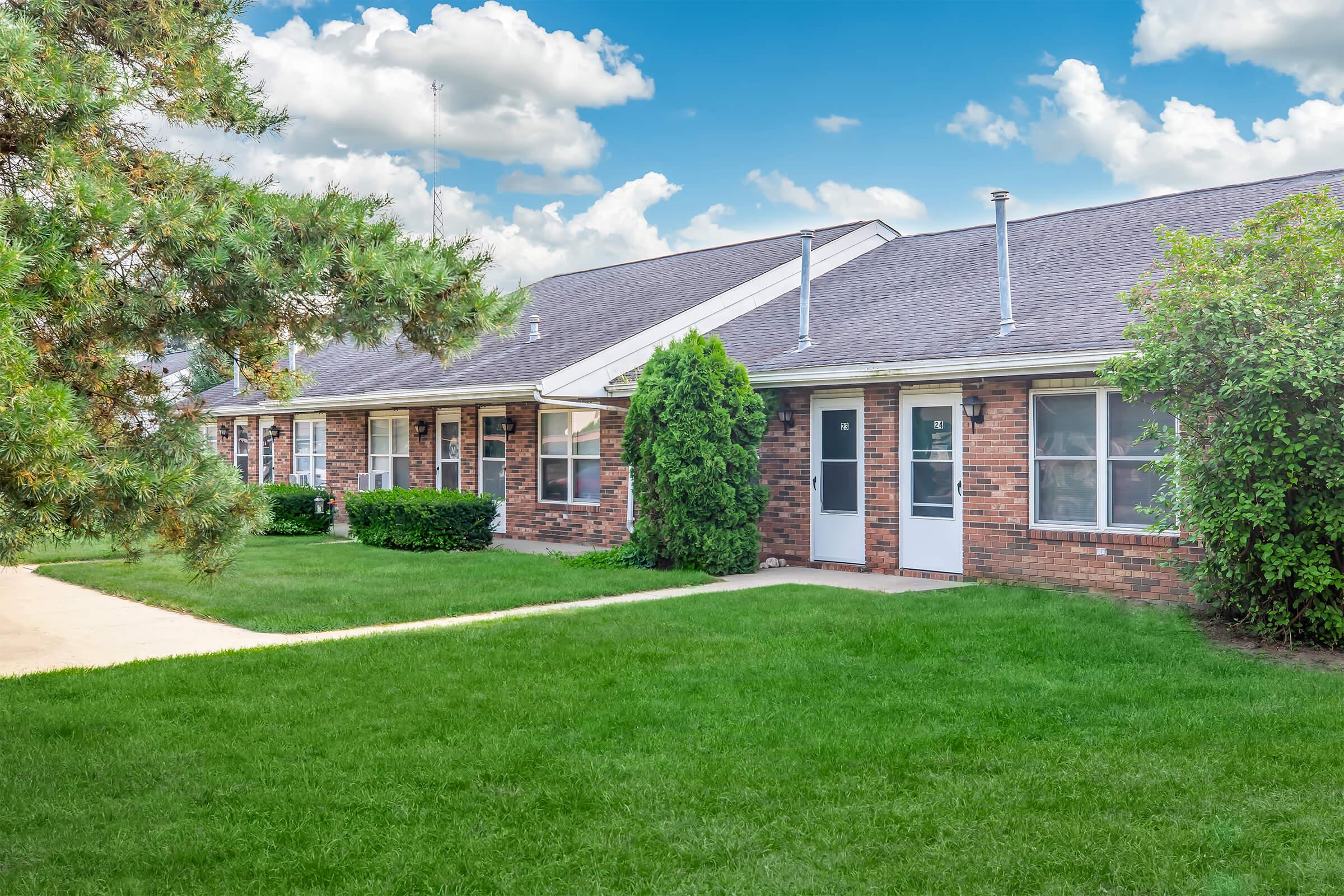 A row of brick apartment units with well-maintained lawns, featuring green shrubs and trees. The sky is partly cloudy, adding brightness to the scene. The pathway leads to the doorways of each unit, showcasing a welcoming residential environment.