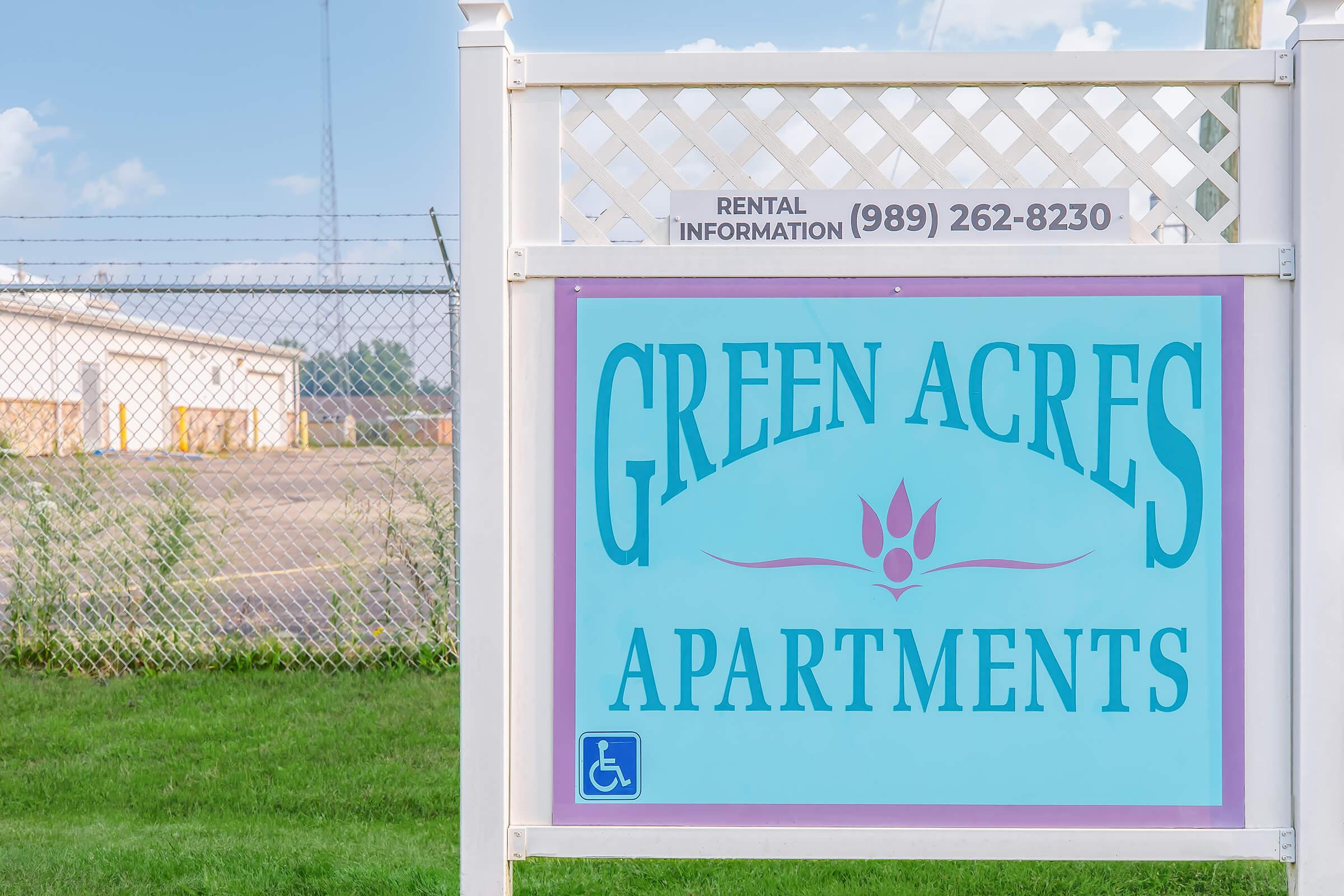 Sign for Green Acres Apartments, featuring a light blue background with purple accents. The sign includes the text "GREEN ACRES APARTMENTS" prominently displayed, along with a rental information phone number, and a wheelchair accessibility symbol. The background features a grassy area and some buildings in the distance.