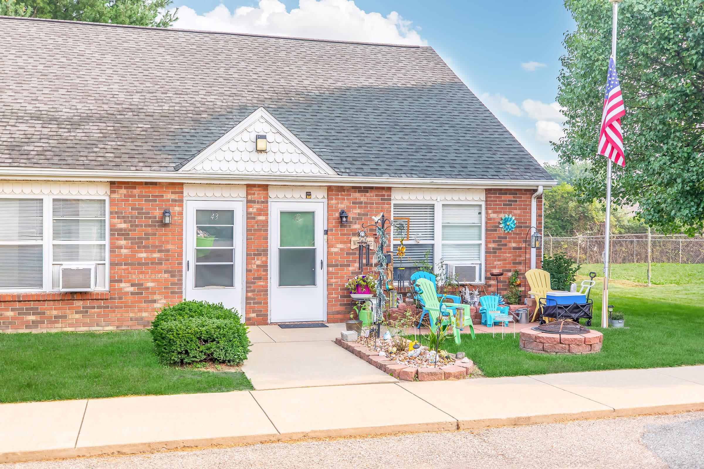A cozy brick-front home featuring a welcoming entrance with a white door and windows, adorned with outdoor seating in bright colors. The front yard has a well-kept lawn and decorative plants, complemented by a flagpole displaying the American flag. Soft clouds float in a blue sky above.
