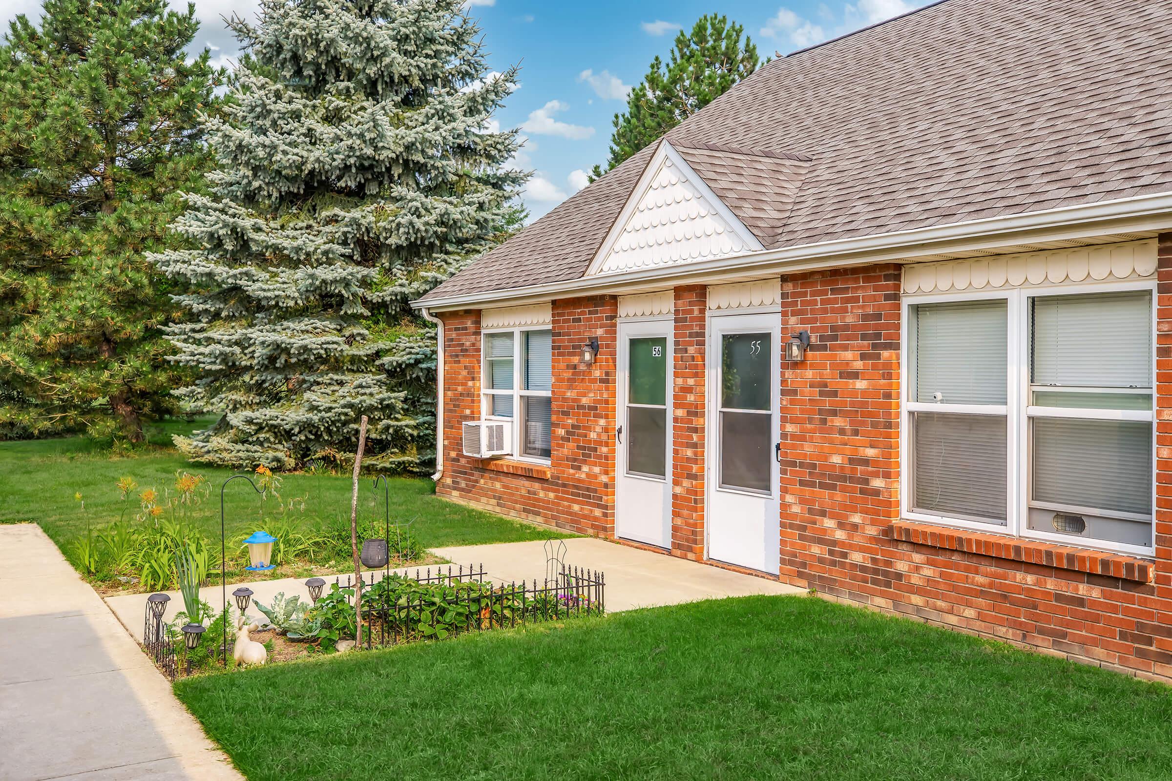 A brick townhouse with two doors, surrounded by a well-maintained lawn and a small garden featuring plants and decorative items. In the background, there are tall pine trees and a clear blue sky with some clouds. The concrete pathway leads to the entrances of the units.