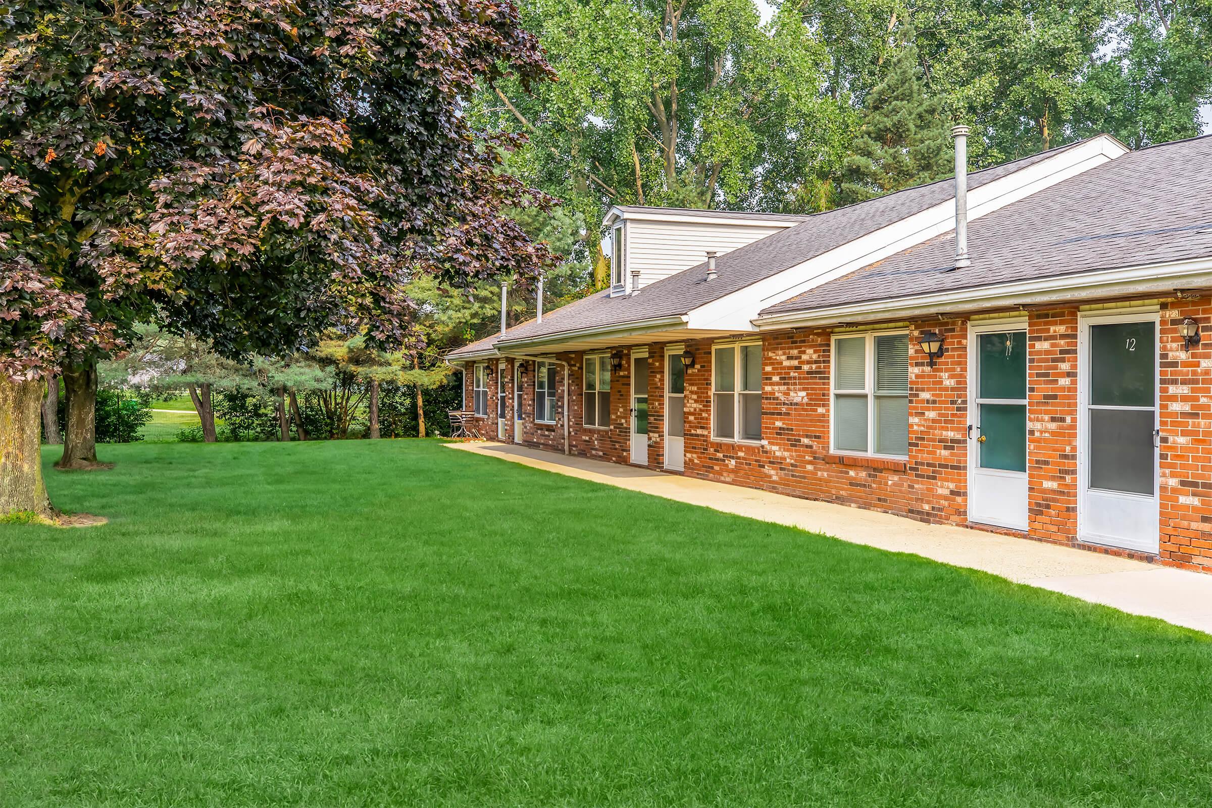 A well-maintained brick building with multiple units, featuring large windows and white doors. The property is surrounded by lush green grass and trees, providing a serene outdoor space. The scene conveys a tidy and inviting atmosphere for residential living.