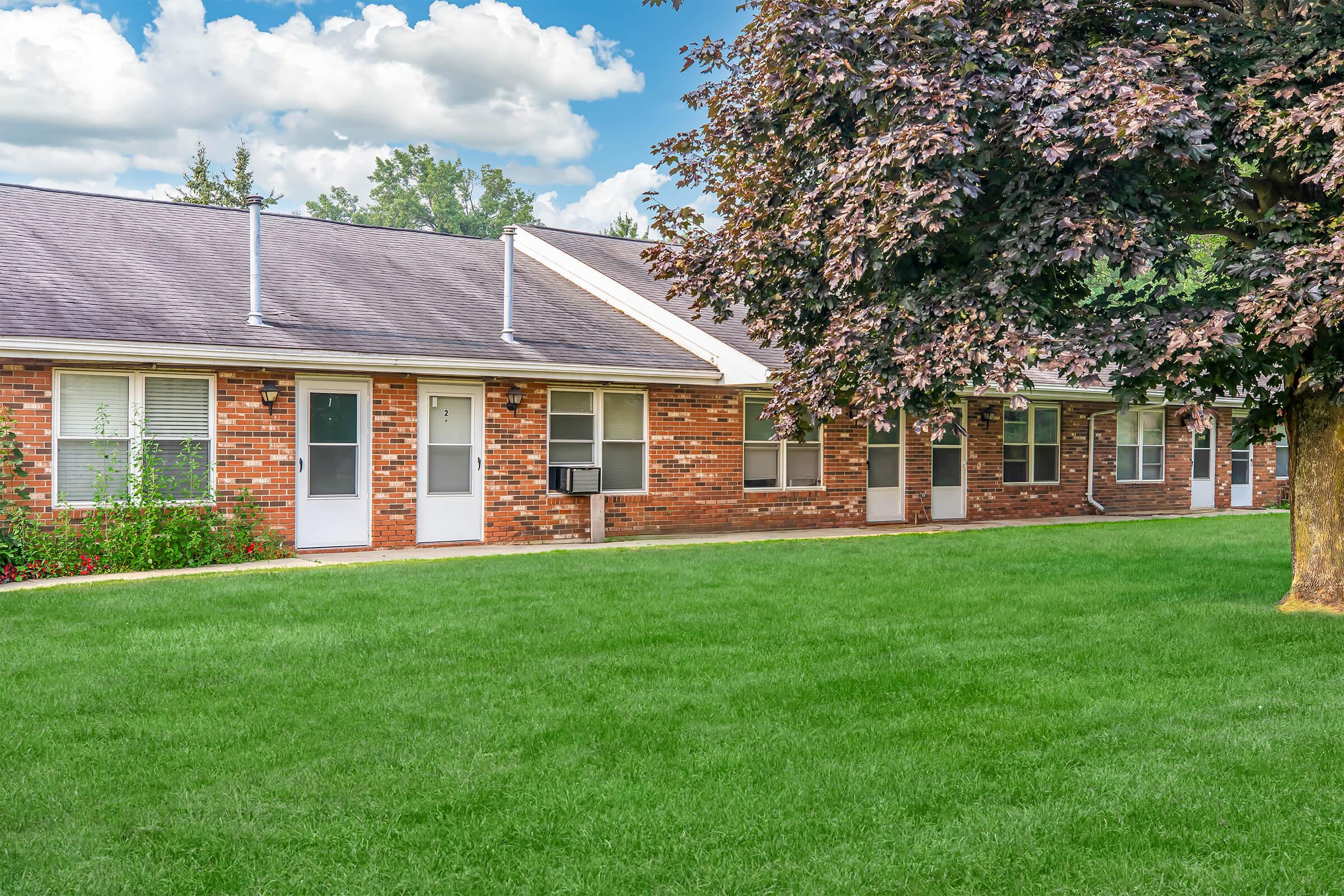 A row of brick apartment units featuring individual entrances, with well-maintained lawns and a large tree nearby. The sky is partly cloudy, and colorful flowers are visible in front of the units, creating a welcoming outdoor space.