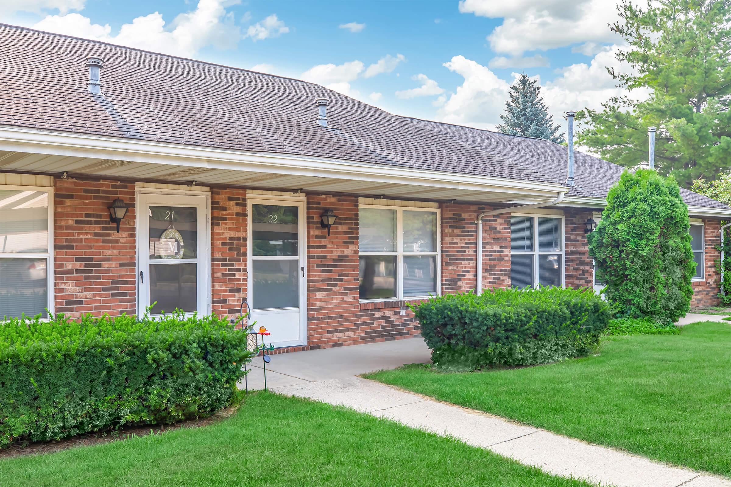Row of brick apartments with freshly mowed green lawns and neatly trimmed hedges. Each unit features a porch with a door framed by windows. Sky is partly cloudy, creating a bright, inviting atmosphere.
