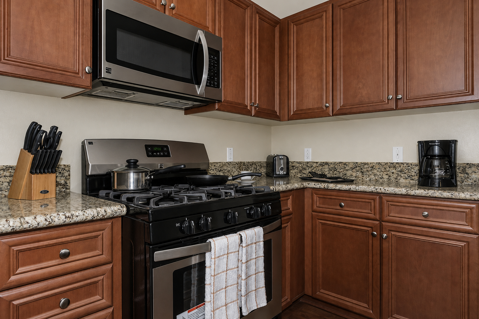 A modern kitchen featuring wooden cabinetry, a stainless steel microwave and gas stove, a granite countertop, and various kitchen appliances including a coffee maker and a knife block. A white dish towel hangs from the stove, adding a homely touch to the space.