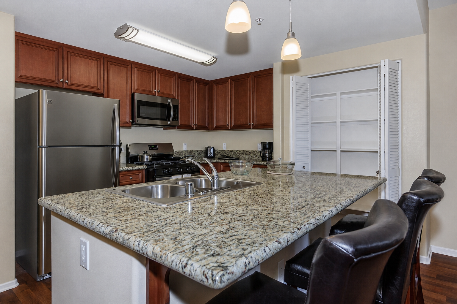 Modern kitchen featuring dark wood cabinets, stainless steel appliances including a refrigerator and microwave, a granite countertop with a double sink, and a small dining area with black barstools. The space is well-lit with pendant lighting and a pantry with sliding doors.