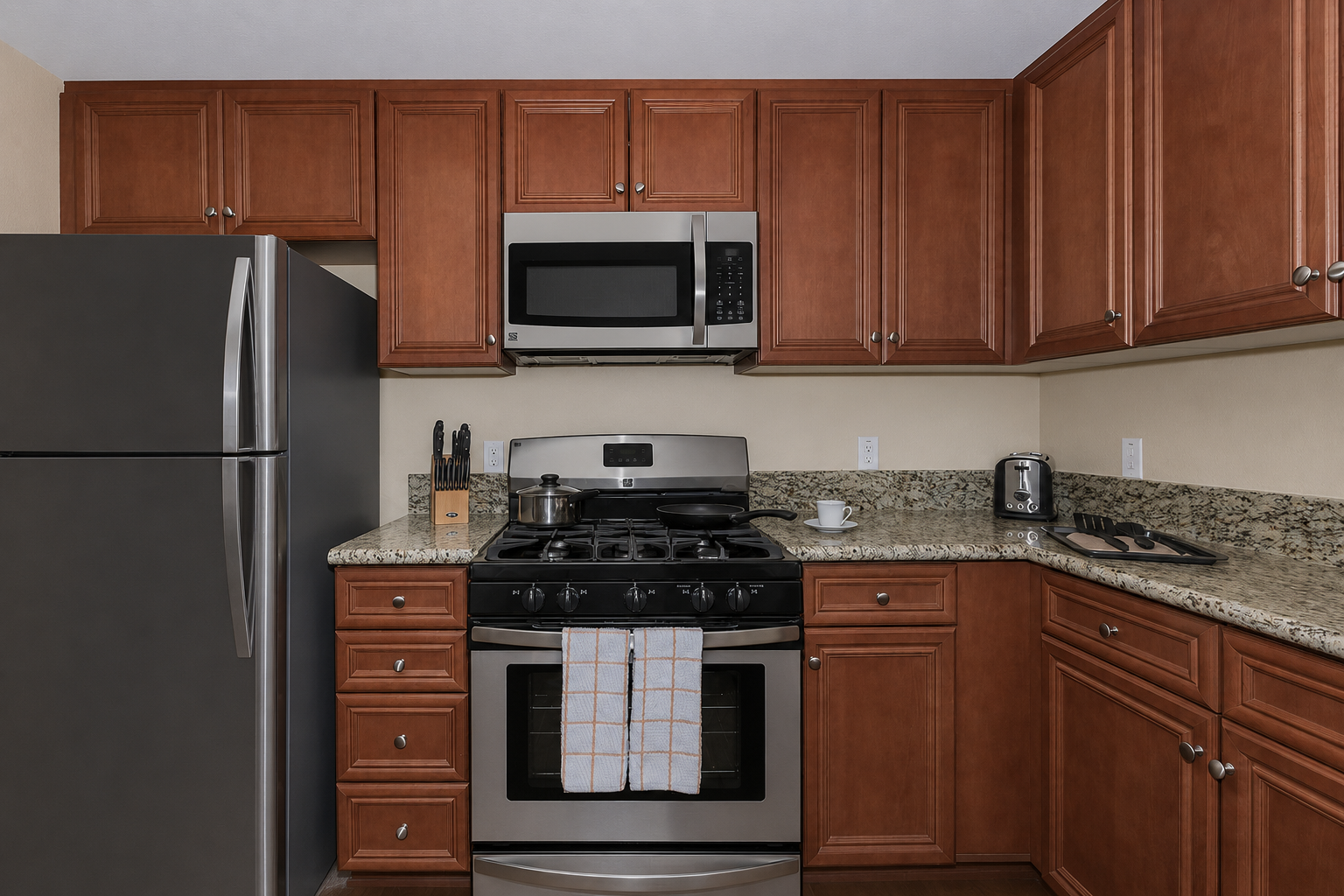 Modern kitchen featuring wooden cabinets, a stainless steel stove and microwave, granite countertops, and a silver refrigerator. Kitchen utensils and a cutting board are visible on the counter, along with a small coffee maker. A dish towel hangs on the stove.