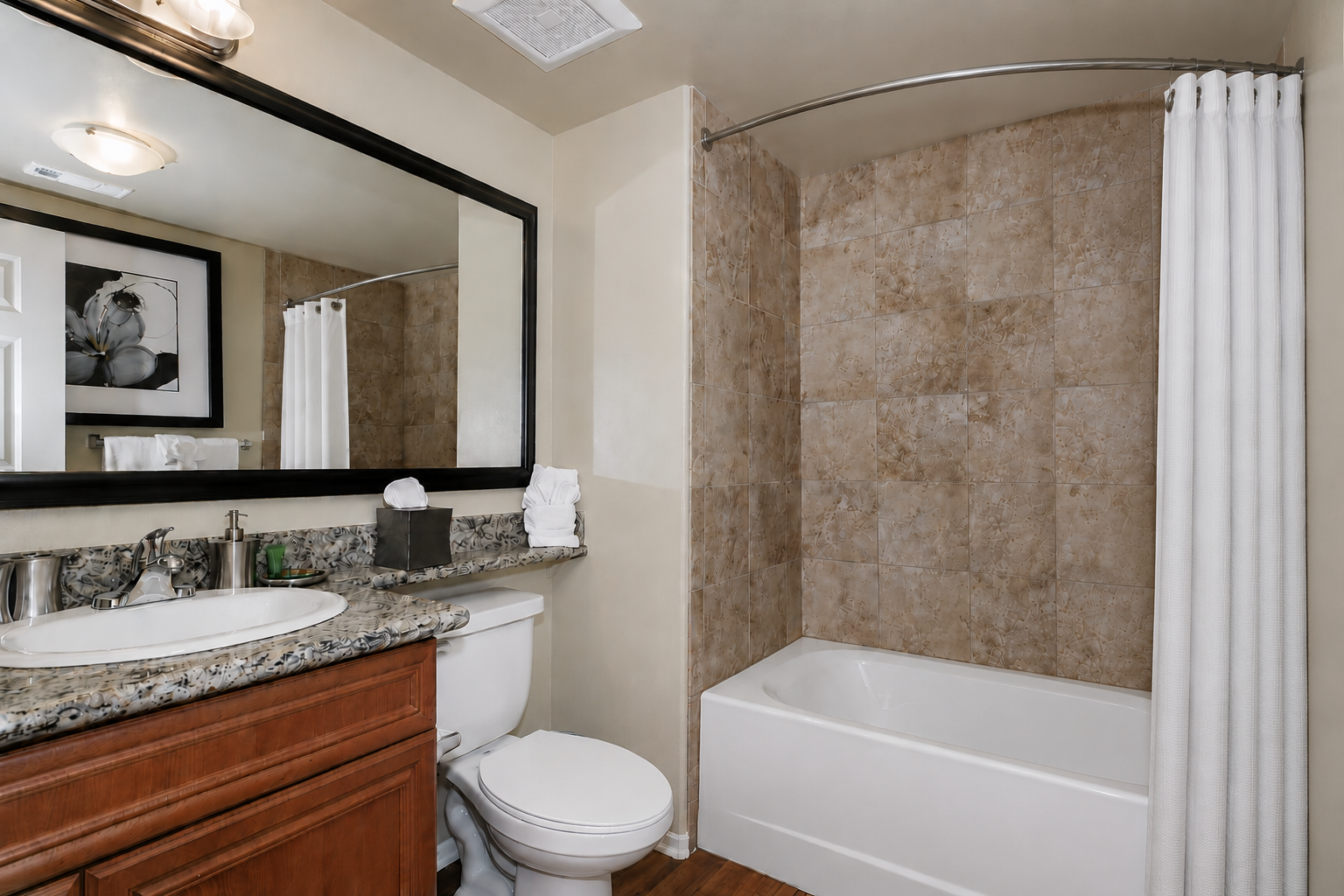A clean and modern bathroom featuring a bathtub with shower curtain, a granite countertop with a sink, a large mirror above the sink, and decorative items. The walls are finished with beige tiles, and the flooring is made of wooden planks. A towel and toiletries are neatly arranged on the countertop.