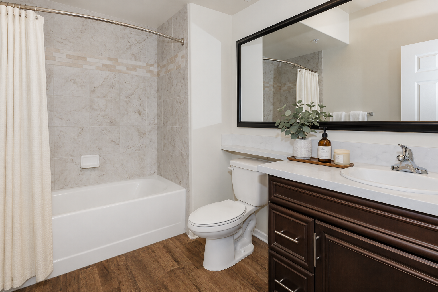 A modern bathroom featuring a white bathtub with a shower curtain, a large mirror above a sink with a marble countertop, a wooden cabinet, and a toilet. The walls have neutral tiles, and there's a touch of greenery on the countertop, creating a clean and inviting atmosphere.