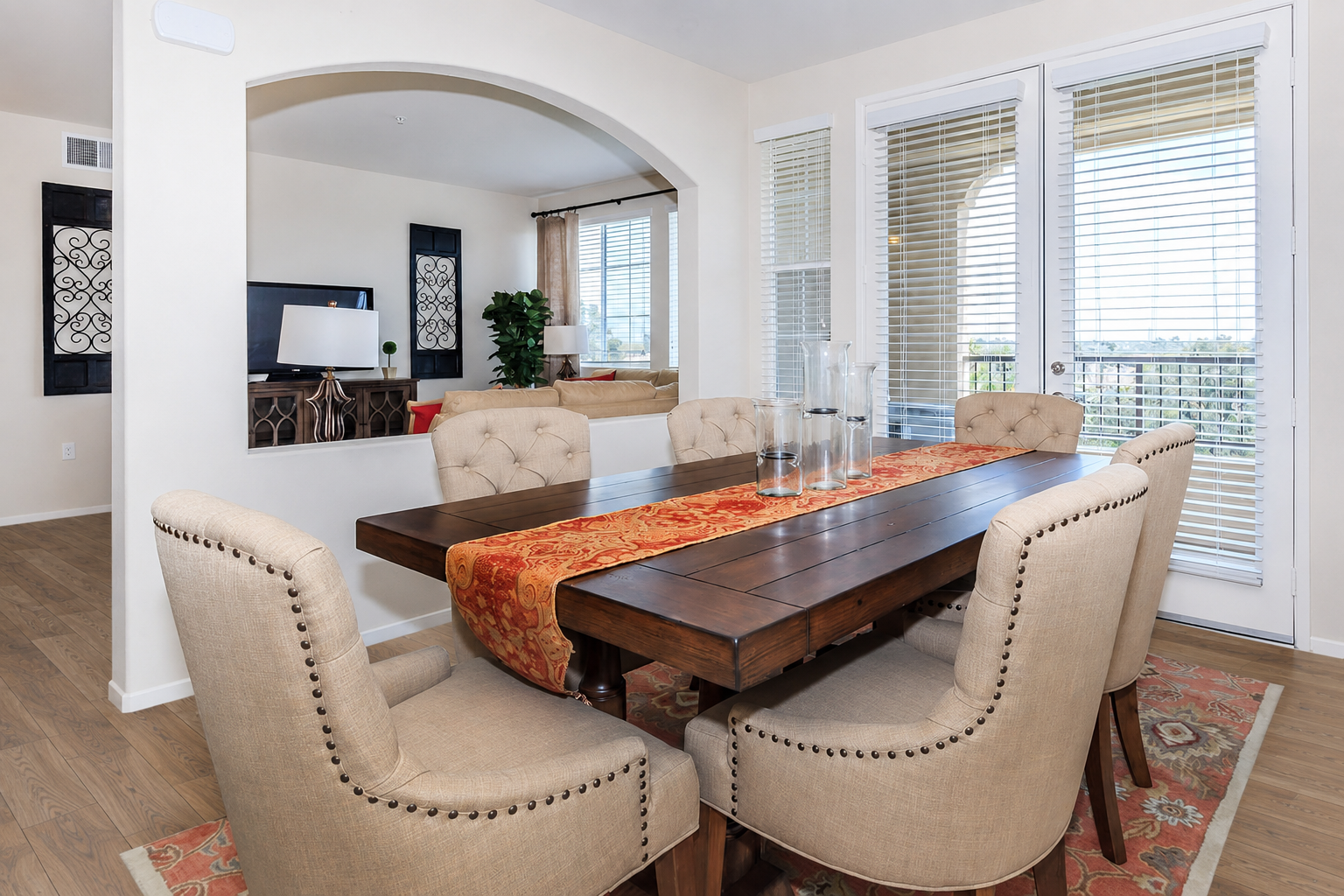 A stylish dining area featuring a wooden table surrounded by beige tufted chairs, with a decorative table runner. The room has an open layout, leading to a living space with large windows draped with blinds, letting in natural light and offering a view of the outdoors.