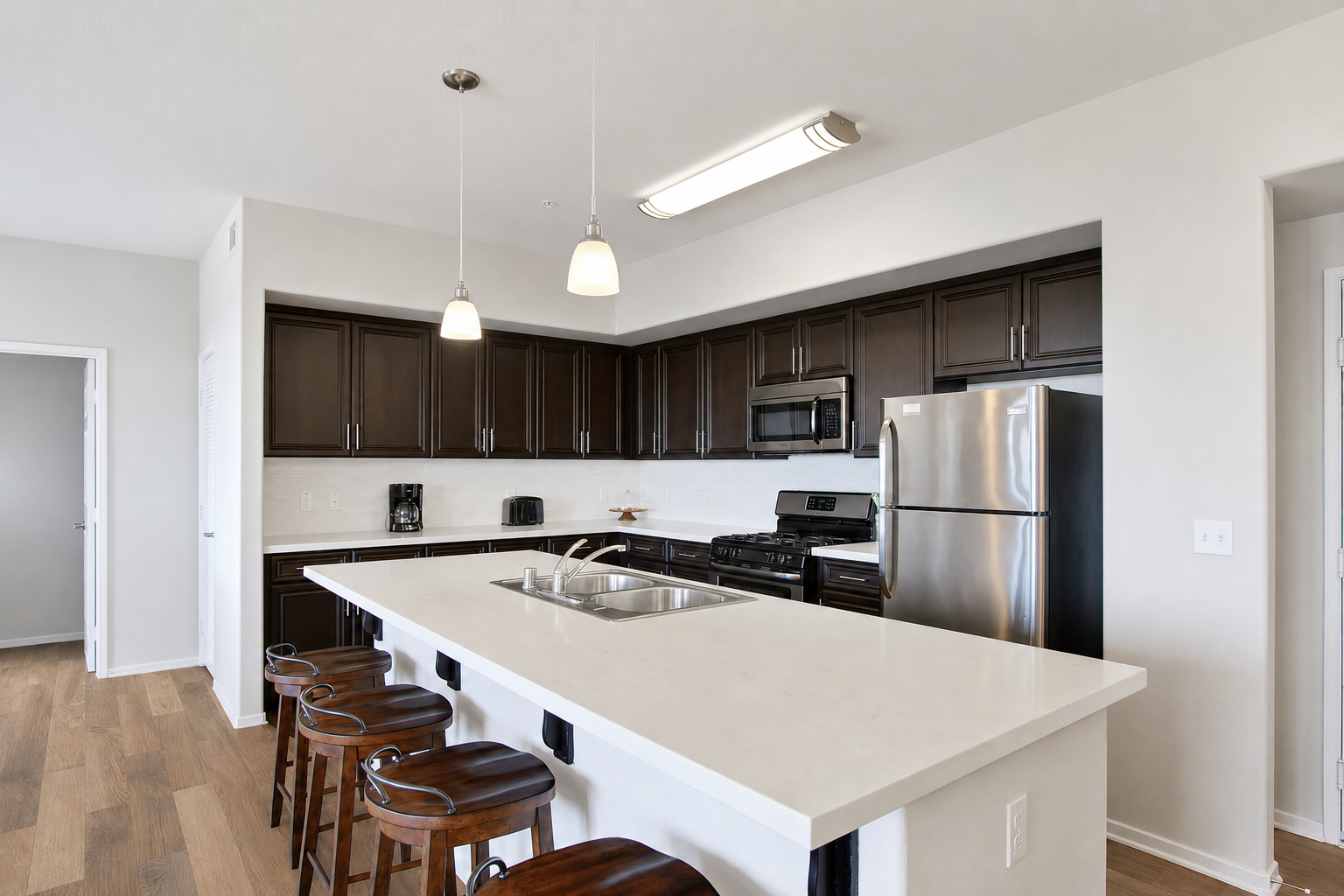 Modern kitchen featuring dark wood cabinets, stainless steel appliances, and quartz countertops. An island bar with four wooden stools is positioned in front, and there are pendant lights above. The space is well-lit with a clean, contemporary design, including a white tile backsplash and laminate flooring.