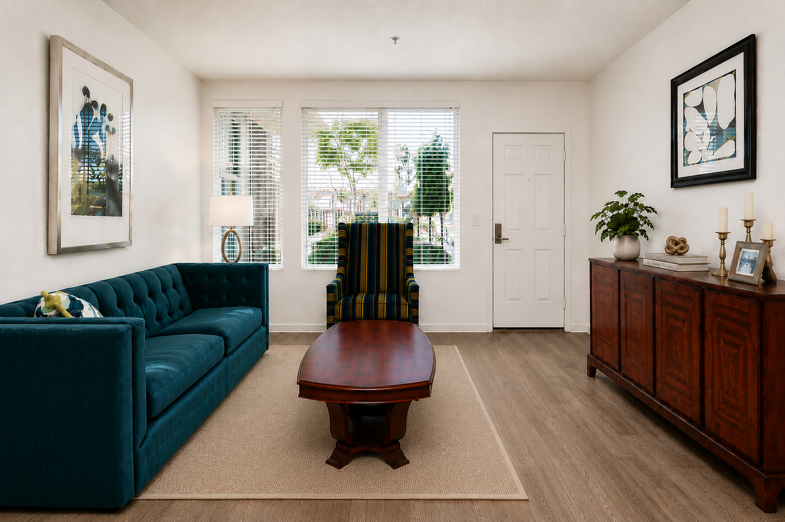 A cozy living room featuring a teal sofa, a wooden coffee table, and a dark wooden sideboard. There are two windows allowing natural light, with greenery visible outside. A tall, striped armchair and decorative items, such as a lamp and a plant, add warmth to the space. Wall art is also present.