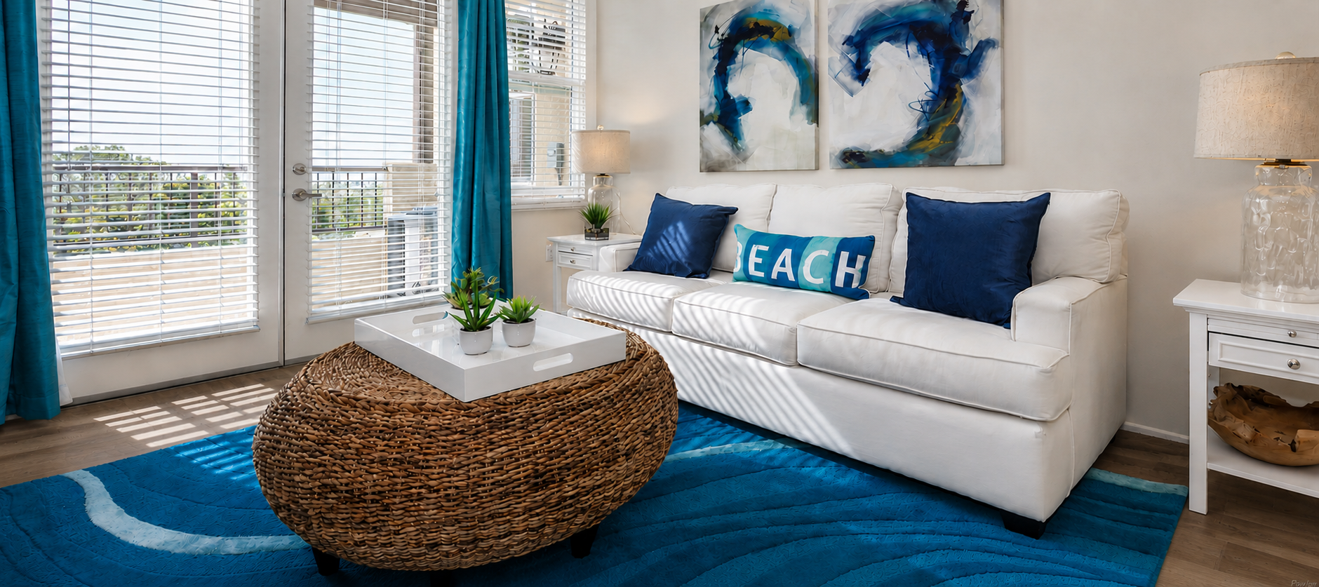 Bright and inviting living room featuring a white sofa adorned with blue accent pillows and a 'BEACH' cushion. A round wicker coffee table sits in front on a textured blue rug. The space is enhanced by large windows with sheer curtains, potted plants, and two lamps, creating a warm, coastal atmosphere.
