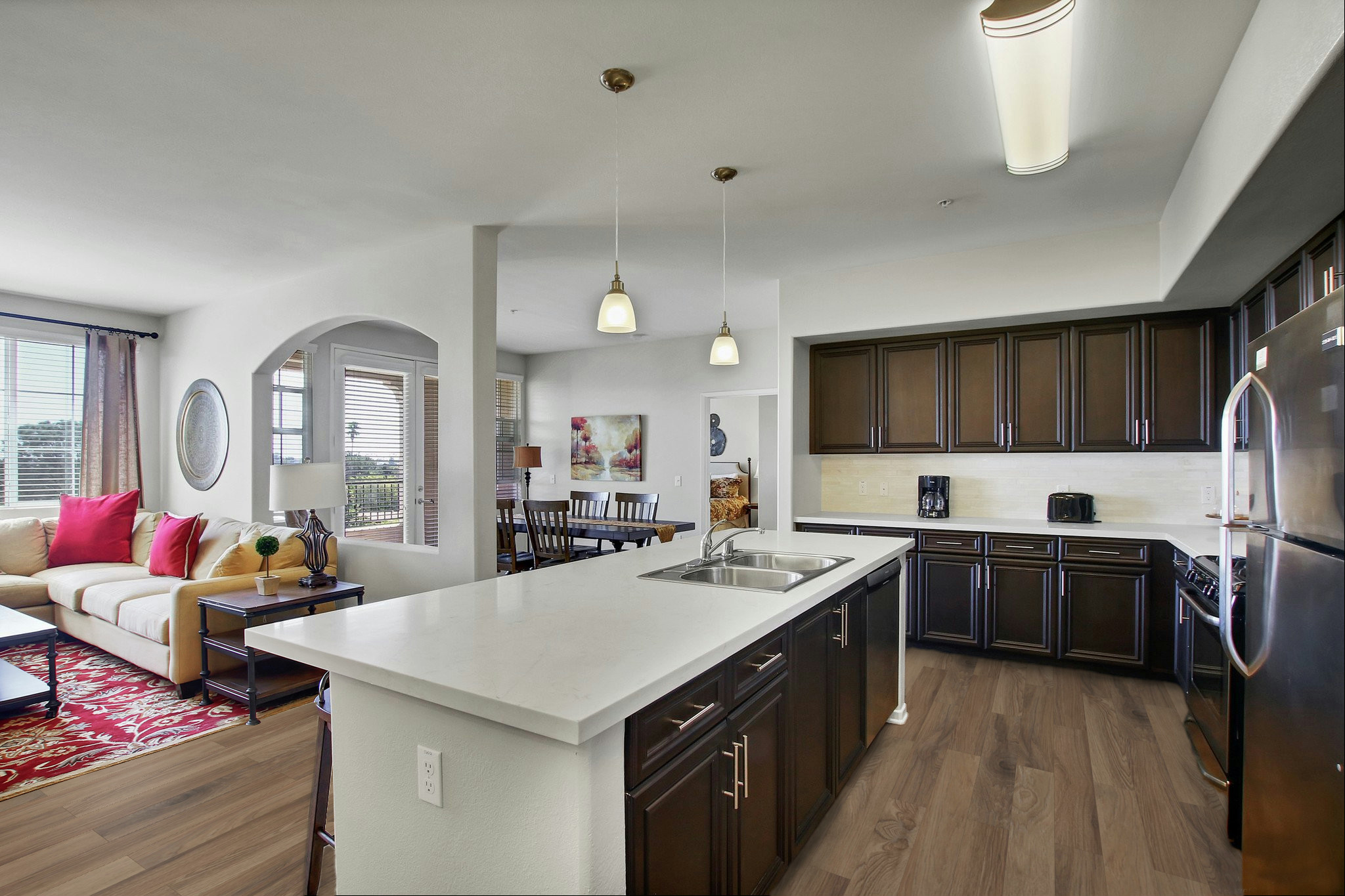 Modern kitchen looking into an open-concept living area. Features dark wood cabinetry, a white countertop with a double sink, and stainless steel appliances. A dining table is visible, along with a comfortable sofa and decorative elements like a rug and wall art, all in a well-lit, spacious environment.