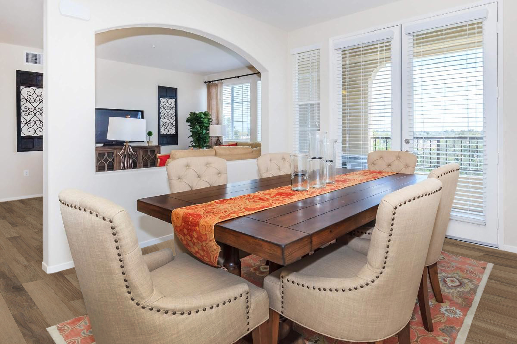 A modern dining room featuring a wooden table surrounded by light-colored tufted chairs. A decorative red table runner is placed on the table. In the background, a living area is visible with a television and plants, and large windows allow natural light to brighten the space.