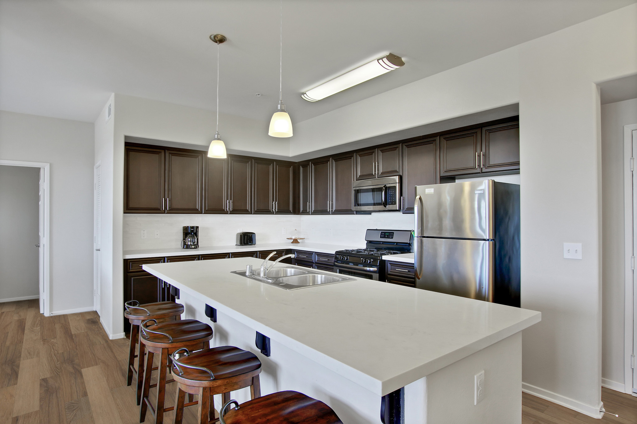 Modern kitchen featuring dark wooden cabinets, stainless steel appliances, and a large island with seating. The space is well-lit with pendant lights and has a clean, open layout, highlighted by light-colored walls and flooring.