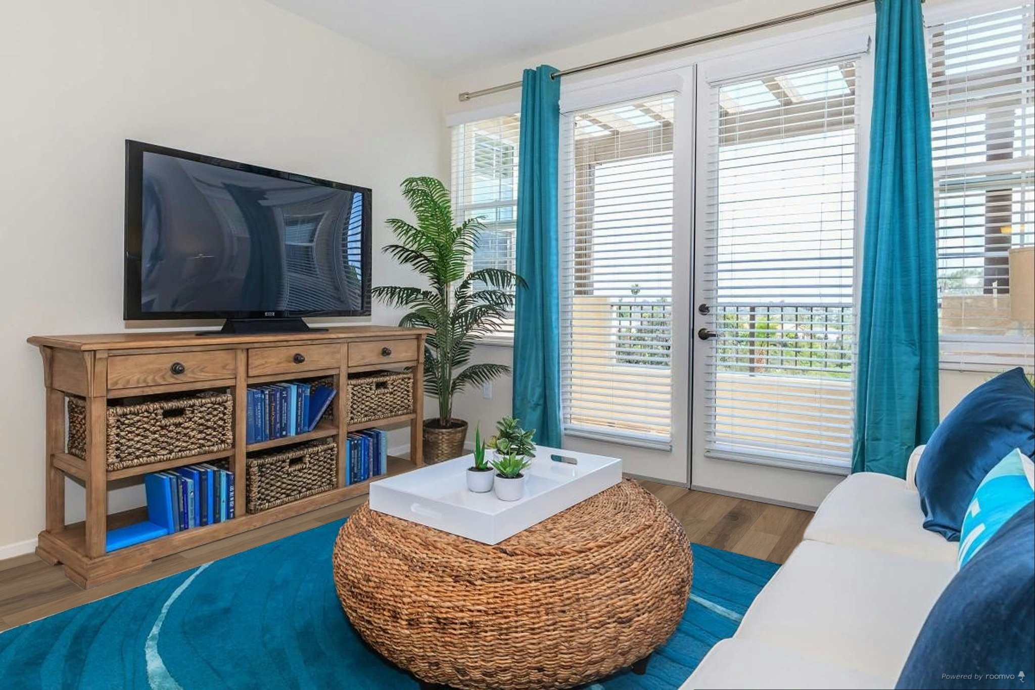 A cozy living room featuring a large TV on a wooden cabinet, a coffee table made of woven material, and a plush white sofa with blue cushions. Light streams in through large sliding glass doors with blinds, complemented by potted plants and a circular blue area rug. Decor elements suggest a relaxed, modern atmosphere.