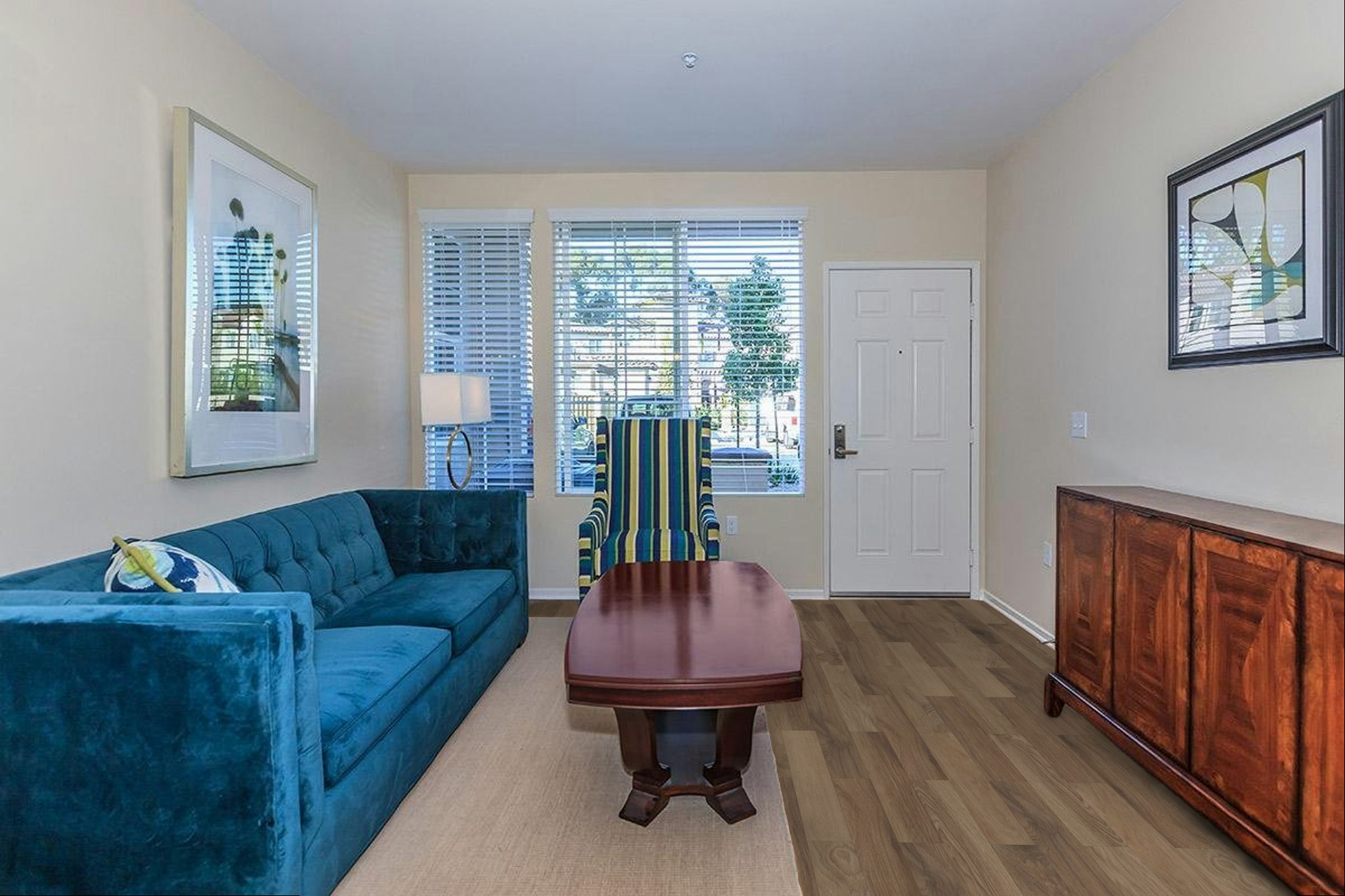 A modern living room featuring a teal couch, a striped armchair, a wooden coffee table, and a sideboard. Bright natural light streams through windows adorned with blinds, illuminating the space's neutral walls and hardwood flooring. Decorative artwork complements the contemporary decor.