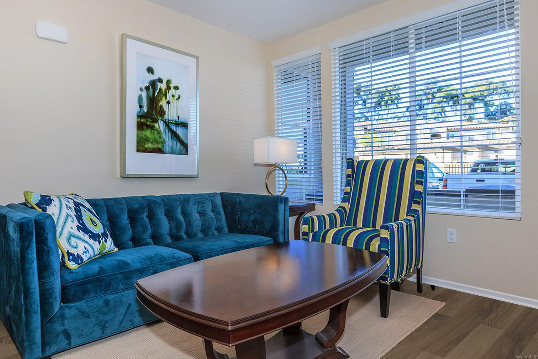 A cozy living room featuring a teal tufted sofa with decorative pillows, a colorful striped armchair, a wooden coffee table, and a lamp. The space is illuminated by natural light coming through large windows with blinds. A framed nature-themed artwork adorns the wall, enhancing the inviting atmosphere.