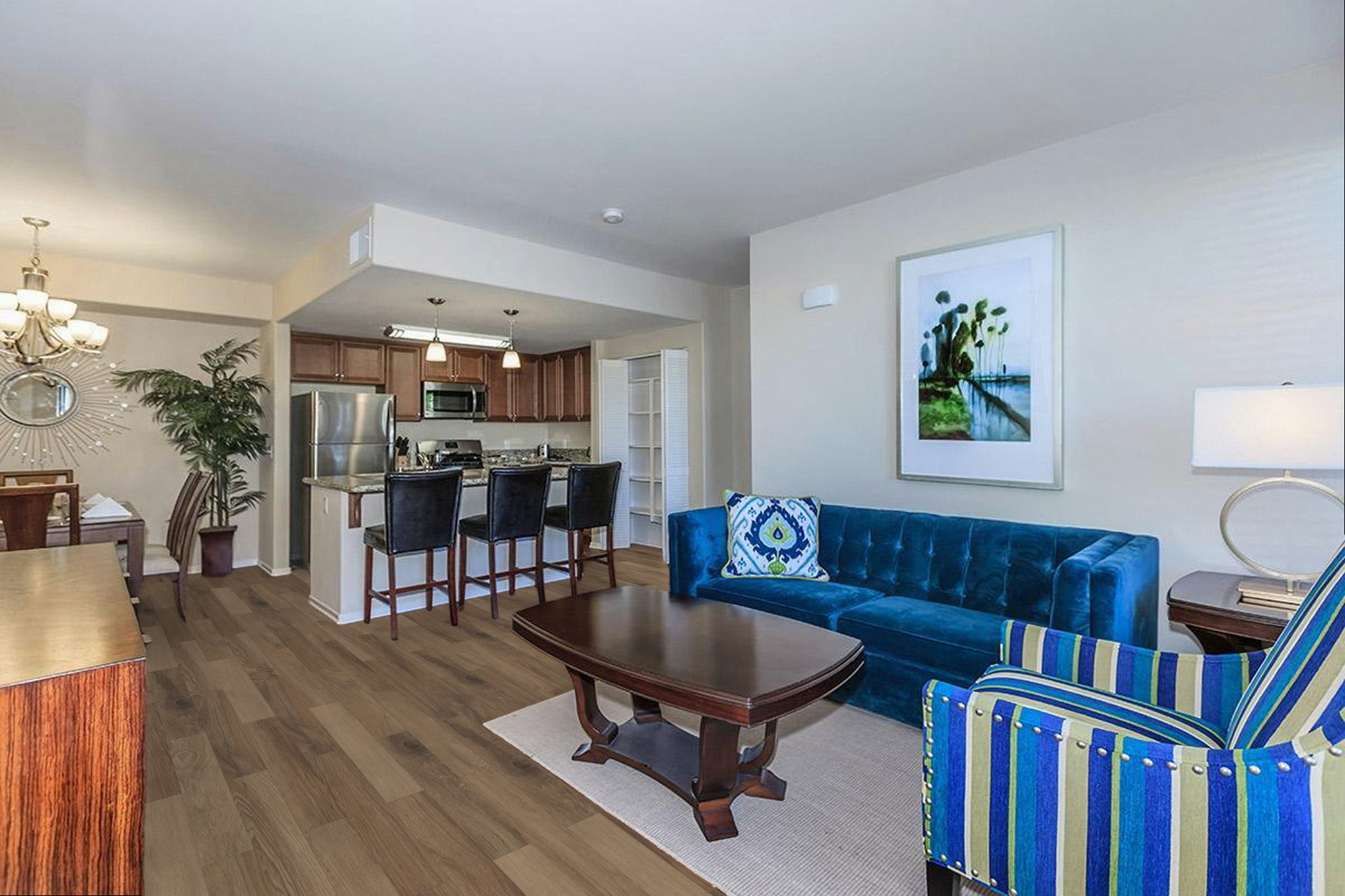 A stylish living room featuring a blue velvet sofa, striped armchair, and a wooden coffee table. The open space includes a dining area and a kitchen in the background, with modern appliances and cabinetry. Decorative artwork and plants add a touch of elegance to the warm, inviting atmosphere.