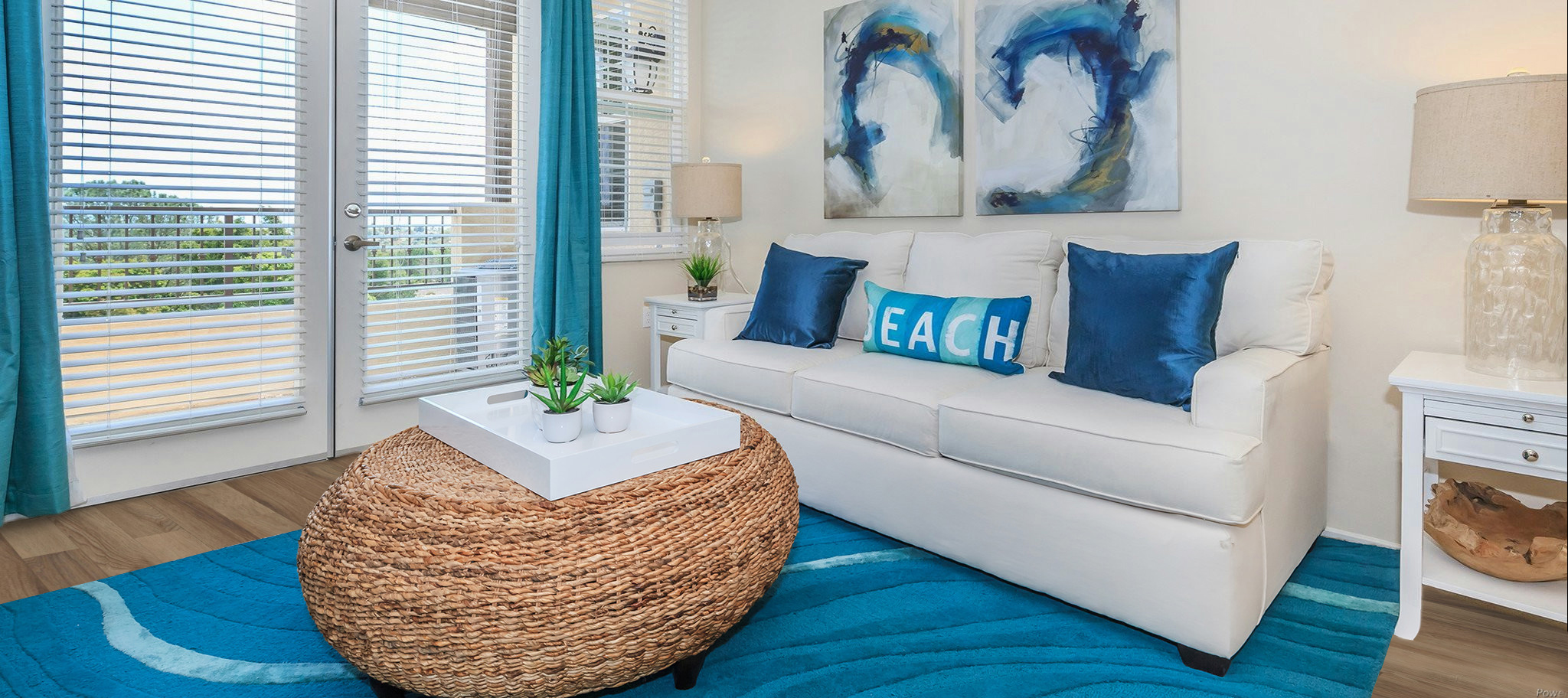 A cozy living room featuring a white sofa with blue throw pillows that say "EACH," a round woven coffee table, and a blue area rug. Large windows with blinds allow natural light to flood in, while abstract wall art adds a touch of color. Decorative plants and lamps complement the inviting atmosphere.