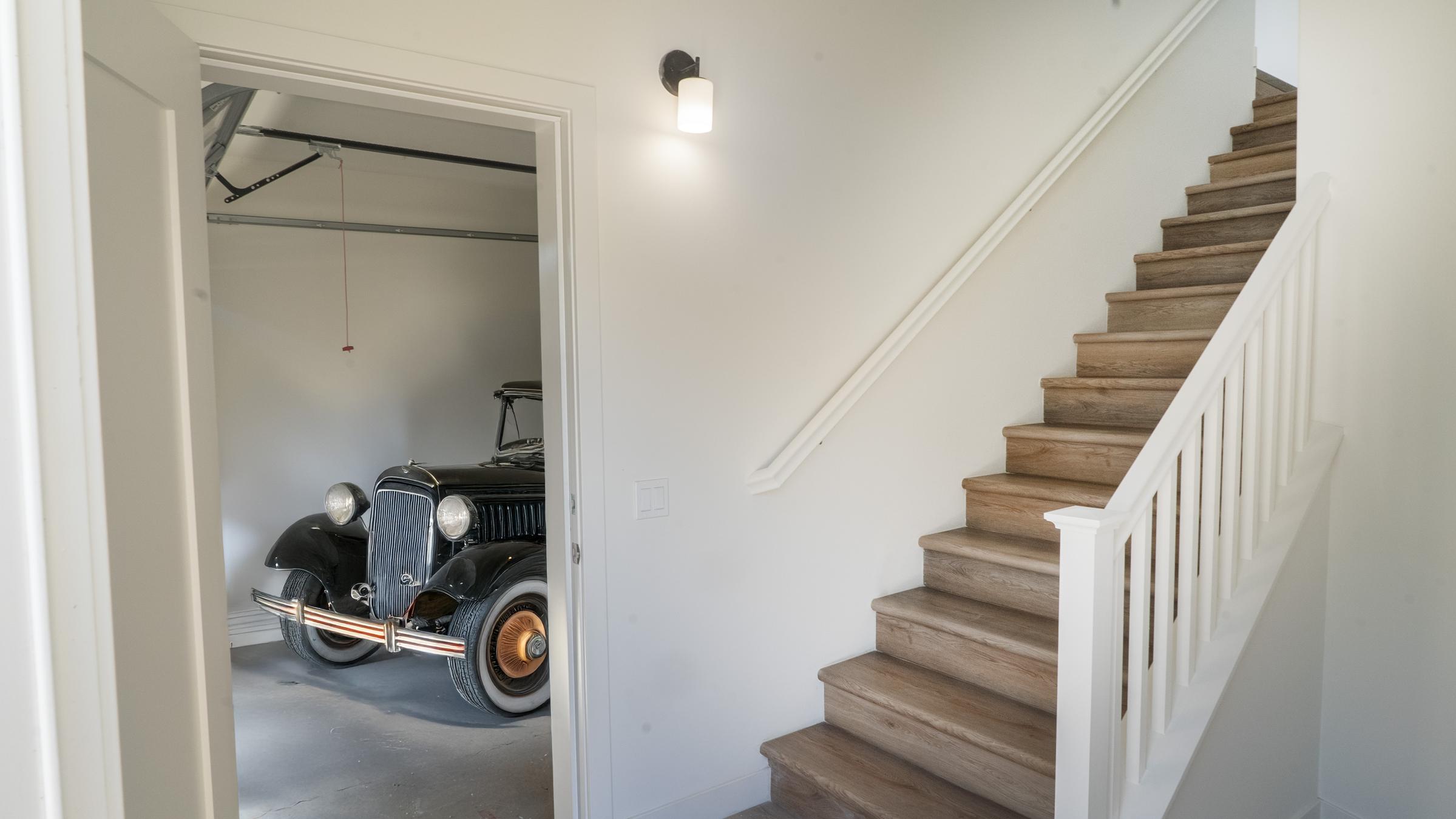 A view of a staircase leading upwards, with bright wooden steps and a white railing. In the background, through an open door, a vintage black car is parked in a garage. The interior features white walls and modern lighting.
