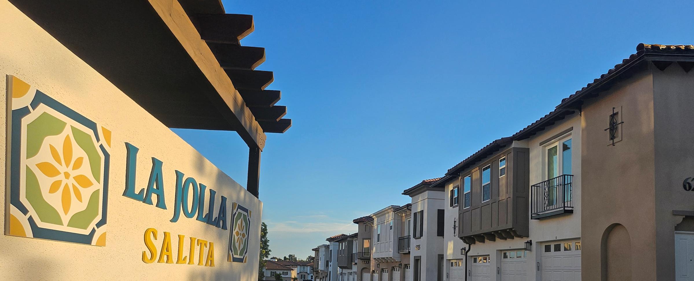 A view of a residential neighborhood featuring modern townhouses. The left side displays a sign that reads "La Jolla Salita," and the architecture includes multiple units with balconies and garages, under a clear blue sky. The scene conveys a welcoming and sunny community atmosphere.