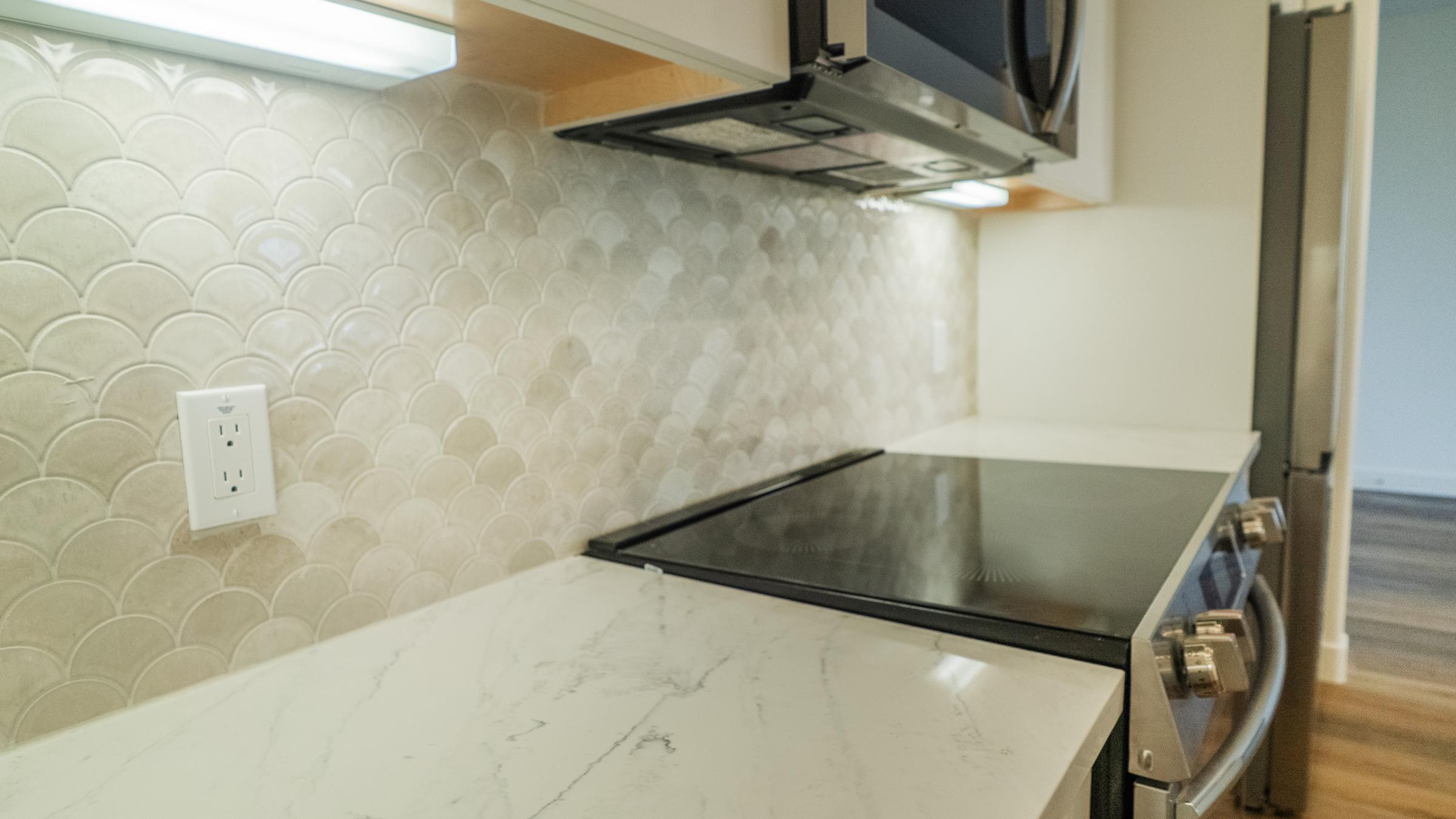 Countertop with a modern design featuring a marble-like surface, a sleek, glass-top stove, and a backsplash made of scalloped tiles. A microwave is mounted above the stove, and there's an electrical outlet visible on the wall next to the countertop. The setting has a clean, contemporary feel.