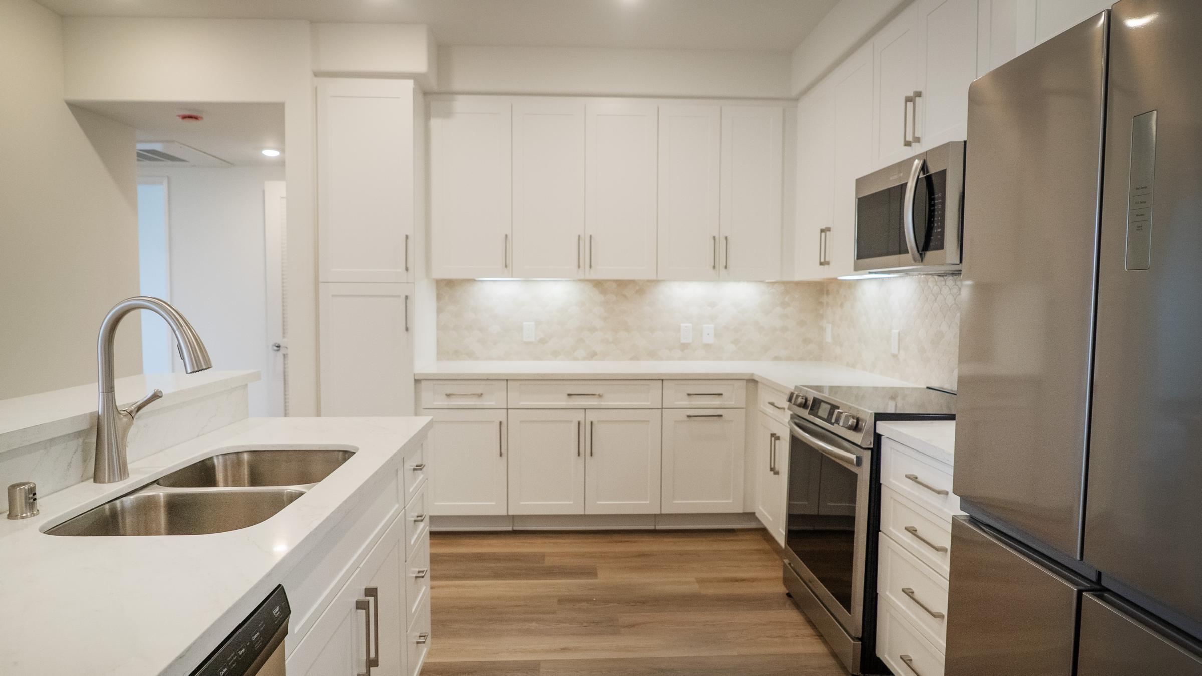 Modern kitchen featuring white cabinetry, stainless steel appliances including a refrigerator, oven, and microwave, a double sink, and a light-colored backsplash. The space has a neutral color palette with hardwood flooring, providing a bright and open atmosphere.