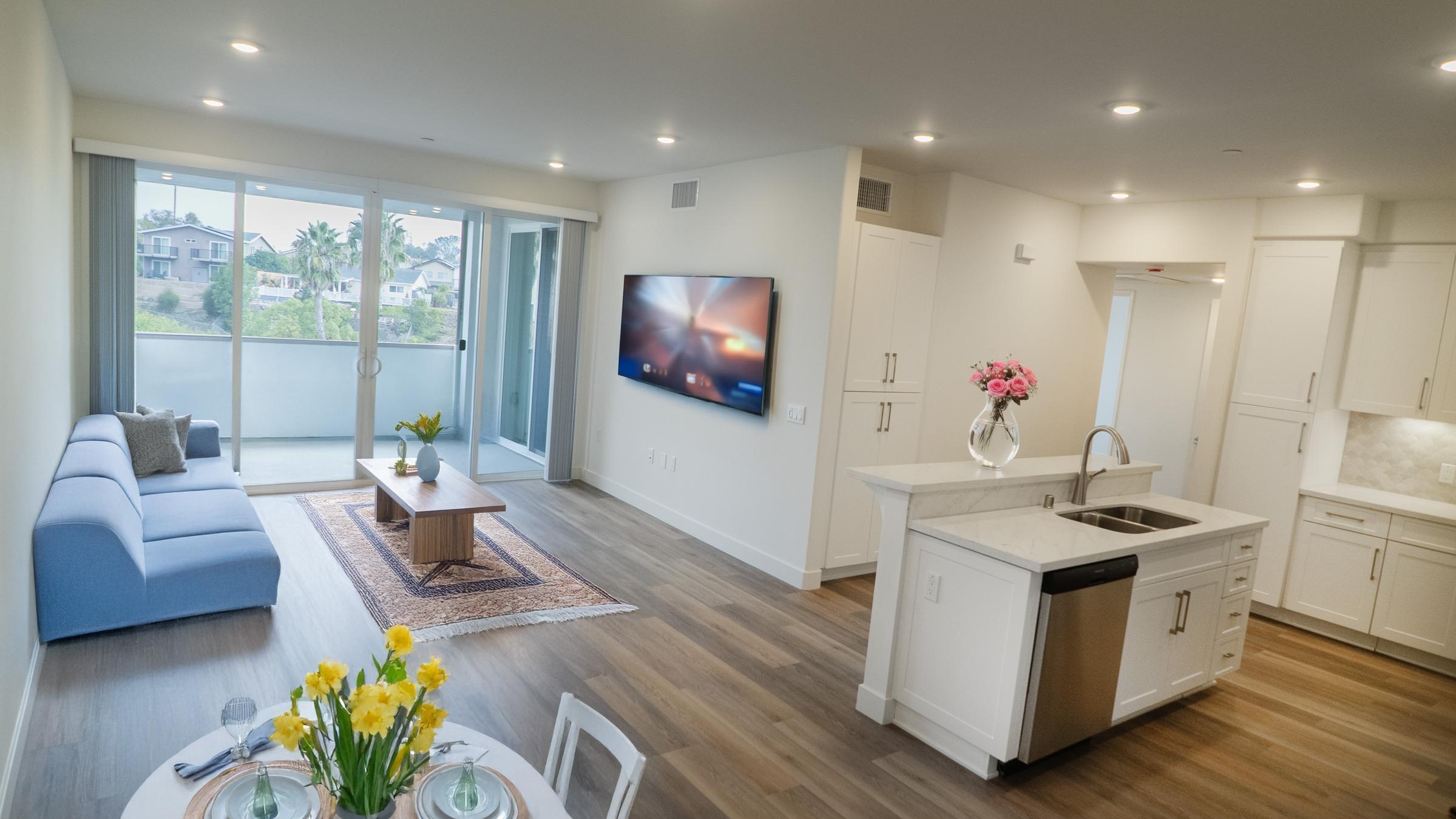 A modern, bright living space featuring a blue sofa, a wooden coffee table, and a dining setup with flower arrangements. A large window offers a view of greenery outside. The kitchen area has sleek white cabinets and stainless steel appliances, with a wall-mounted TV in the living area.