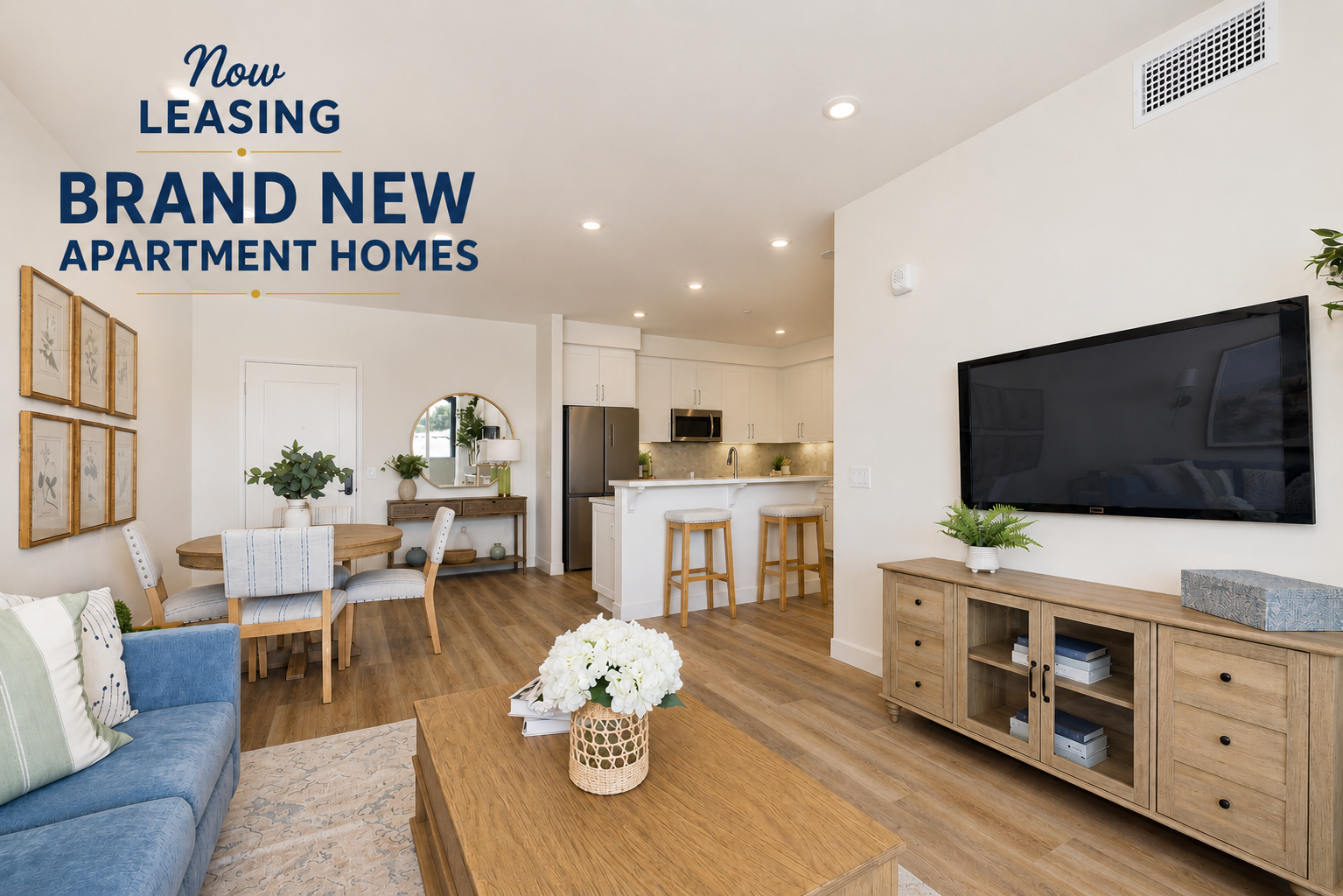 A modern living room featuring a blue sofa, wooden coffee table with flowers, a TV on a wooden media console, and a dining area with a round table and chairs. The kitchen is visible in the background, showing a clean, contemporary design. Text overlay indicates "Now Leasing Brand New Apartment Homes."