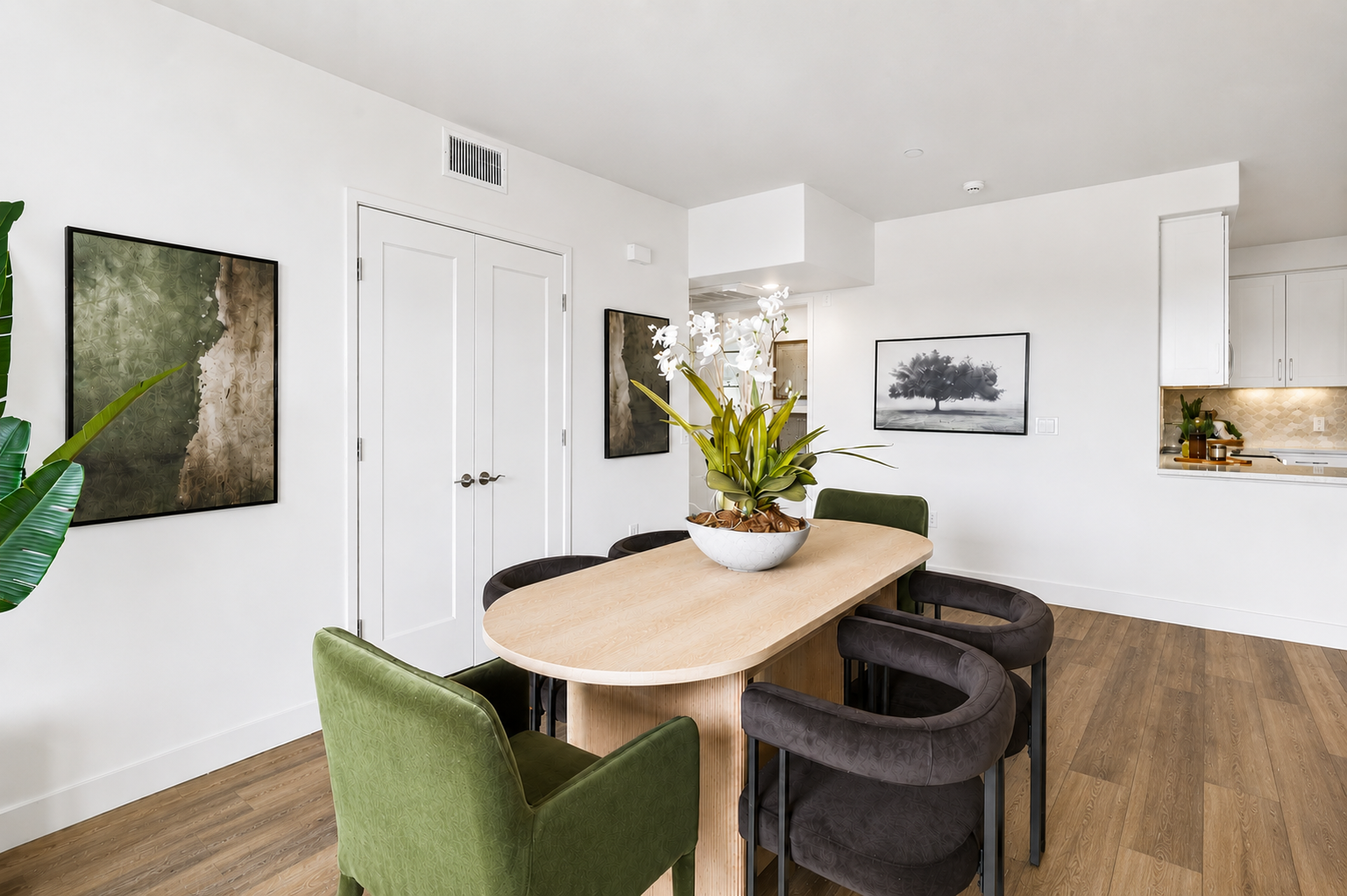 A modern dining area featuring a wooden table surrounded by green and dark chairs. Potted orchids center on the table. Abstract art pieces hang on the light-colored walls, and large windows allow natural light to illuminate the space, enhancing its contemporary aesthetic. A glimpse of a kitchen area can be seen in the background.