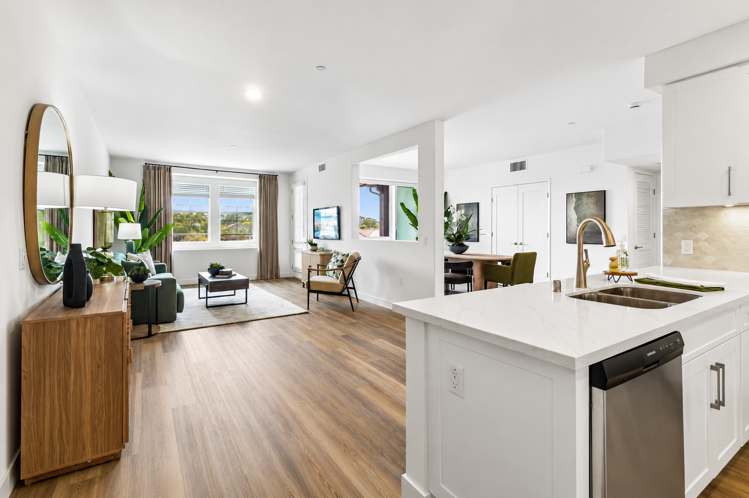 A modern interior space featuring an open-concept living area with hardwood floors. There’s a spacious kitchen with white cabinetry and a stainless steel sink, a dining area, and a cozy living room with plants and a large window providing natural light. A circular mirror and stylish furniture add to the decor.