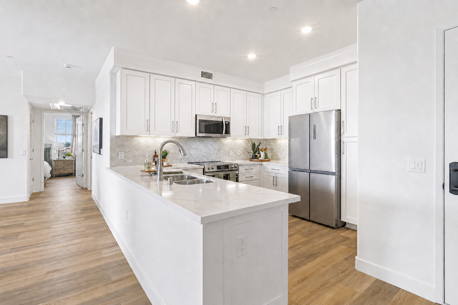 Bright modern kitchen featuring white cabinetry, stainless steel appliances, and a large island with a marble countertop. The open layout includes a view into a living area and natural light from large windows. The flooring is light wood, adding warmth to the space.