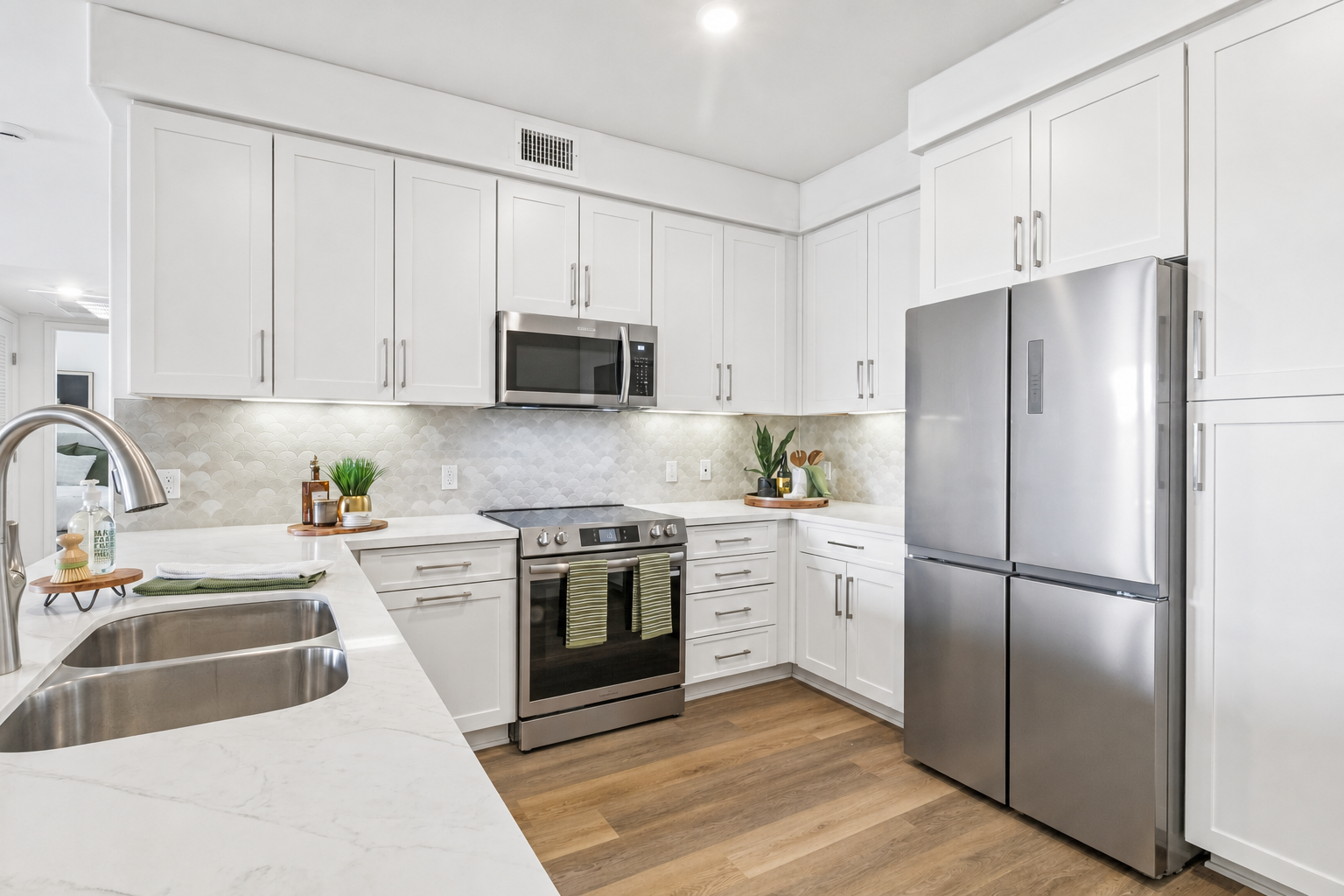 Bright modern kitchen featuring white cabinetry, a stainless steel refrigerator, a built-in oven, and a microwave. The countertops are made of light marble, and there is a double sink with a sleek faucet. The backsplash is a neutral tile, and the space is well-lit with recessed lighting. Small plants and kitchen accessories are displayed on the counters.