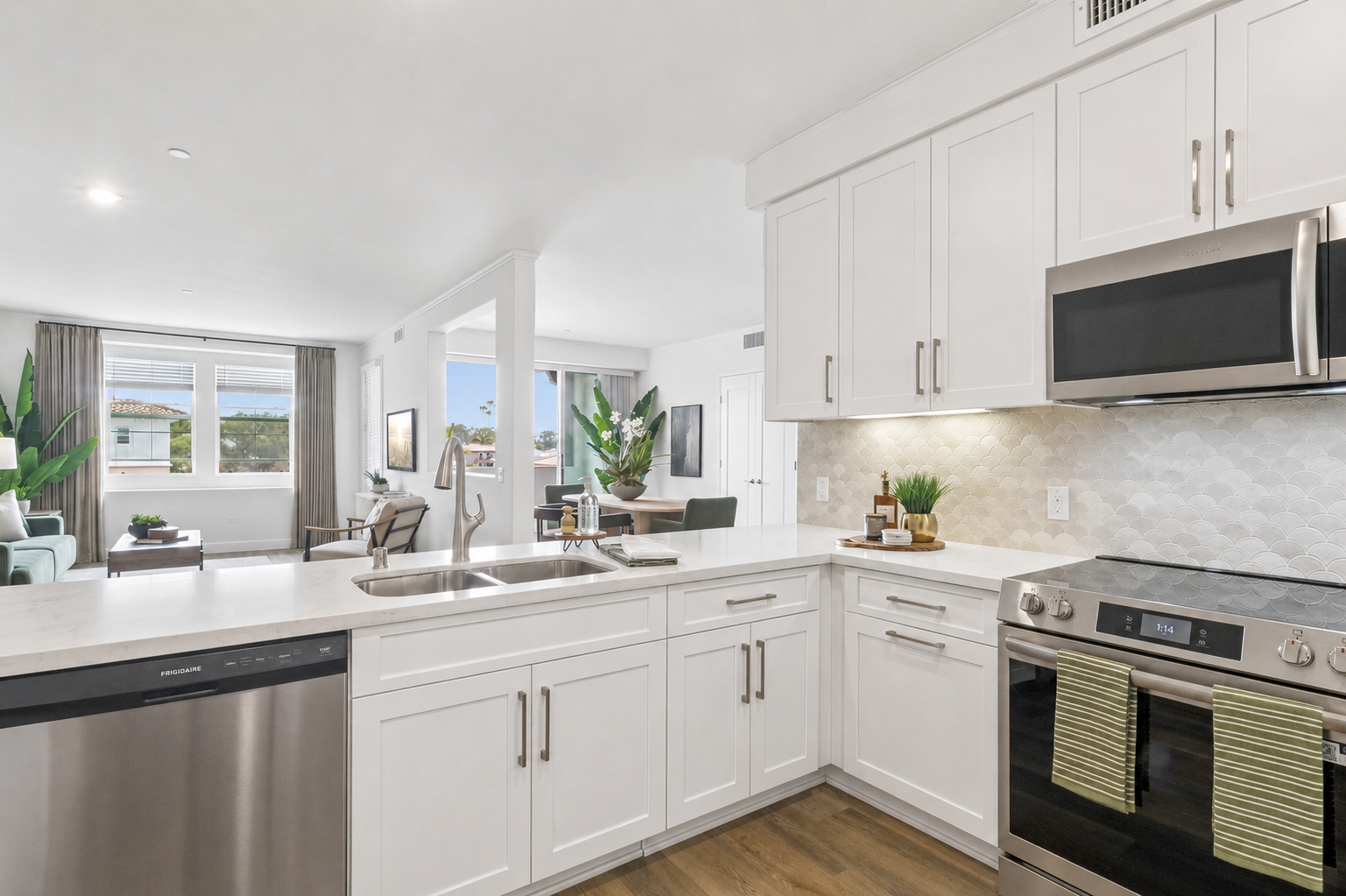 Modern kitchen featuring white cabinetry, stainless steel appliances, and a large countertop. Includes a sink, dishwasher, and an oven. Natural light fills the space, highlighting the stylish backsplash and decorative plants. The open layout connects to a living area with contemporary furnishings in the background.
