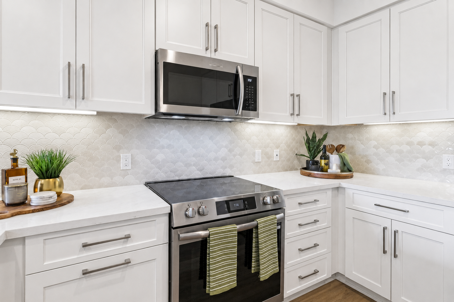 Modern kitchen with white cabinetry, a stainless steel microwave above a sleek stove. The counter features a wooden cutting board with utensils and a plant, accented by green-striped towels hanging from the stove. The backsplash is made of light-colored, scalloped tiles, creating a clean and bright atmosphere.