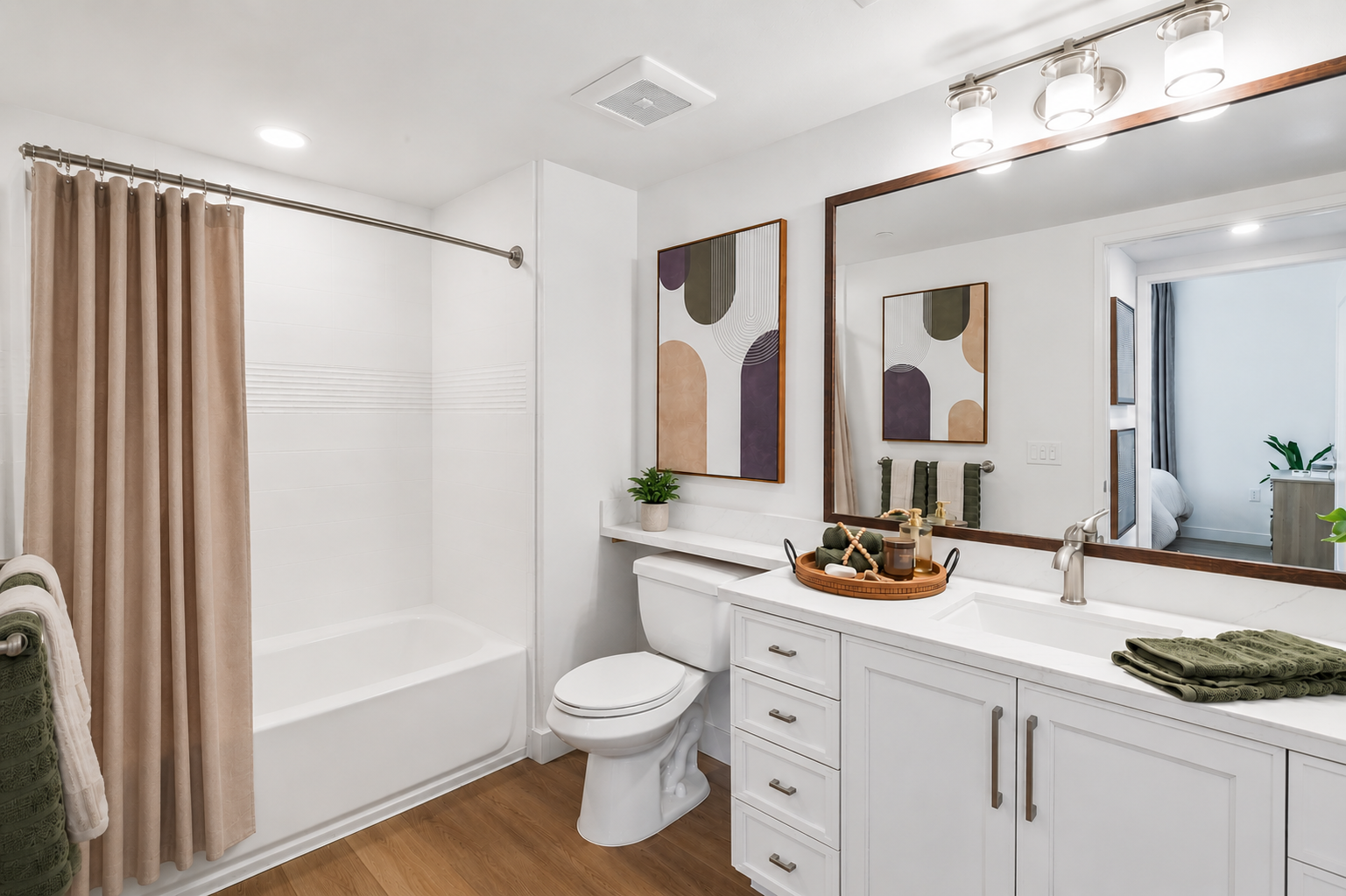 A modern bathroom with a white bathtub and beige shower curtain. The vanity features a white countertop with a sink, a mirror above, and decorative items including a plant and a wooden tray. Neutral-toned artwork hangs on the walls. Green towels are displayed on a rack, and the flooring is wood.