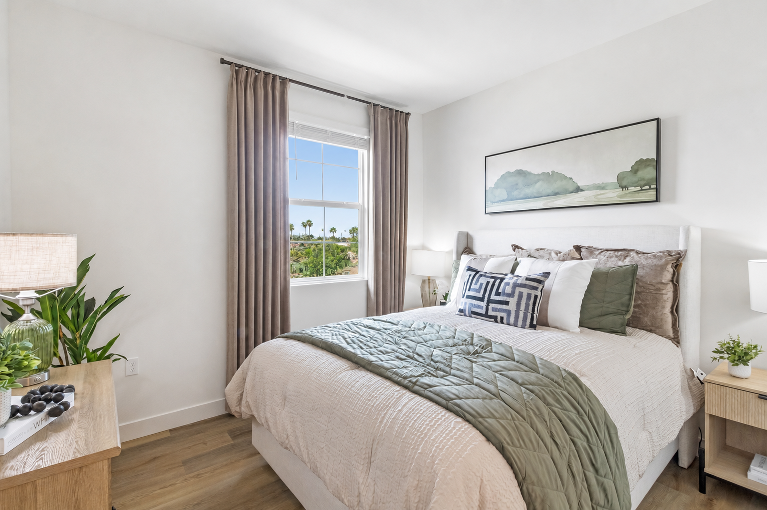 A cozy bedroom featuring a light-colored bed with decorative pillows, a green quilt, and a wooden nightstand with a lamp. A large window provides natural light, showing a view of palm trees outside. Minimalistic decor enhances the peaceful ambiance of the space.