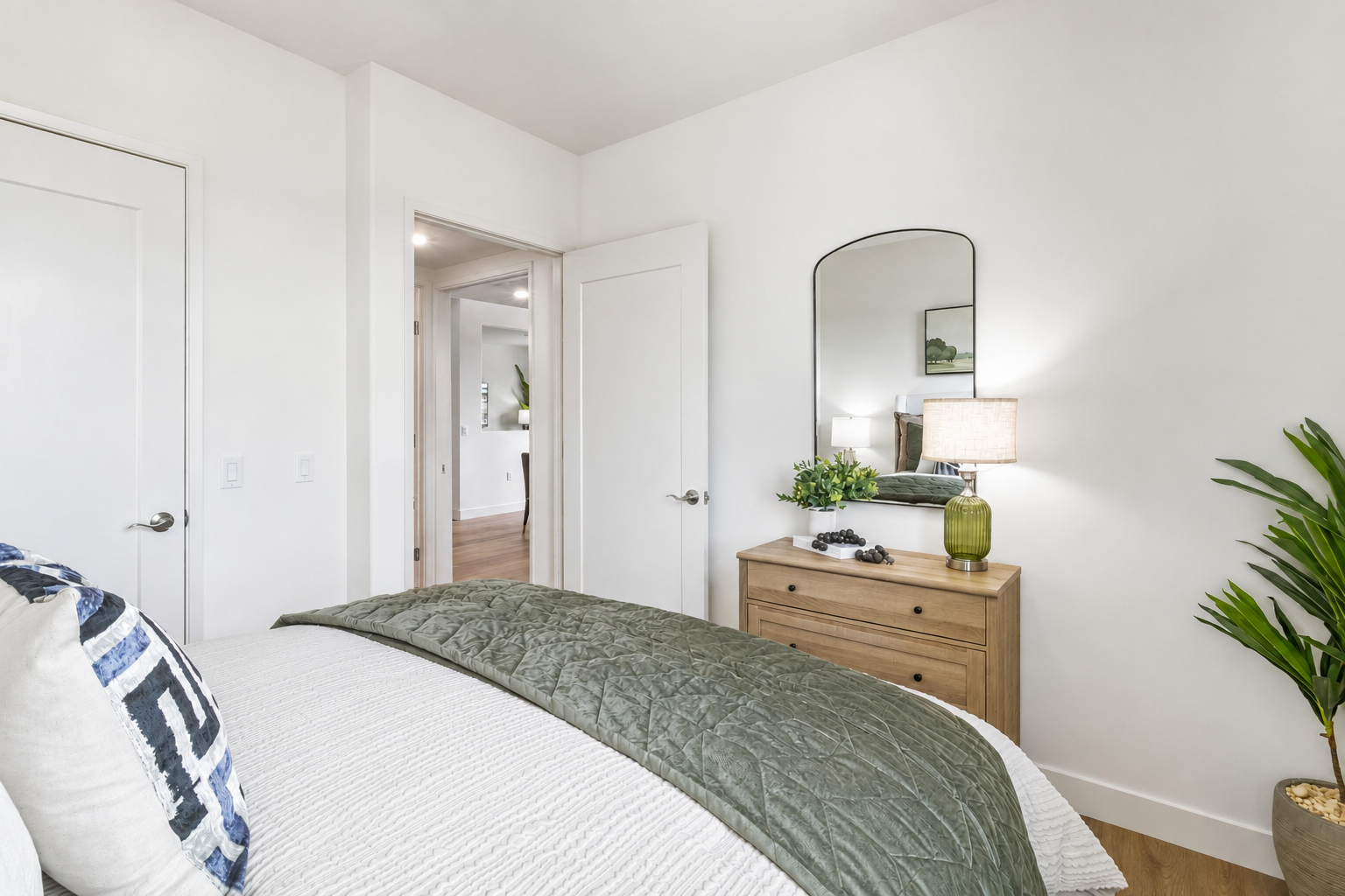 A modern bedroom featuring a bed with a green quilt, a wooden dresser with a lamp, and a decorative mirror. A plant in the corner adds a touch of greenery. The walls are painted white, creating a bright and airy atmosphere. A doorway leads to another room, enhancing the spacious feel.