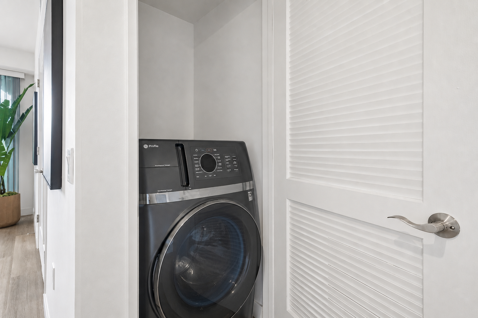 A modern washing machine in a small laundry nook, located in a compact space with light-colored walls. There's a partially closed louvered door and a decorative green plant in a pot visible in the background, enhancing the contemporary feel of the interior.
