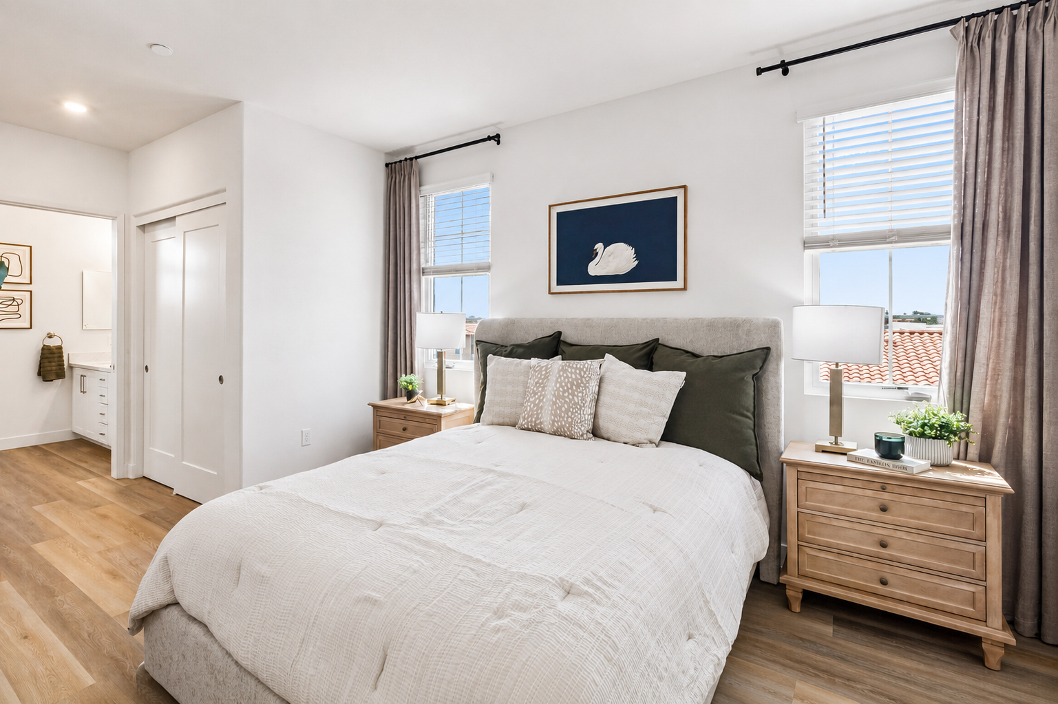 A cozy bedroom featuring a light-colored bed with decorative pillows, two wooden nightstands with lamps, and a framed artwork of a swan on the wall. Natural light streams in through two windows with curtains, illuminating a light wood floor and minimalist decor.