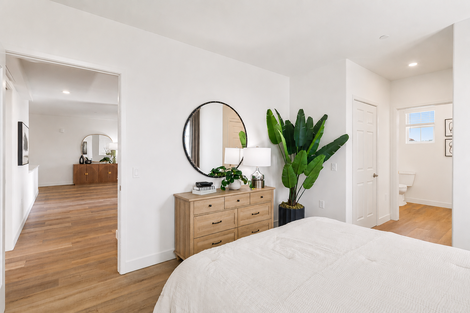 A cozy bedroom featuring a large bed with a light-colored comforter. There's a wooden dresser with a round mirror above it, a lamp, and a potted plant beside the bed. The room leads to a hallway, with a glimpse of a separate room in the background, all boasting warm wooden flooring and bright, airy lighting.