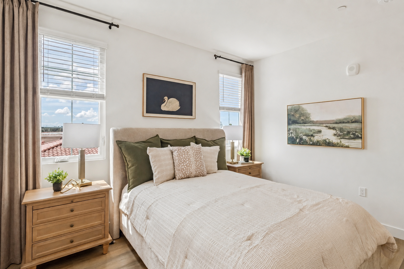 A cozy bedroom featuring a plush bed with decorative pillows, two bedside tables with lamps, and large windows allowing natural light. The walls are adorned with two framed artworks, and the overall color scheme includes soft neutrals and greens, creating a tranquil and inviting atmosphere.