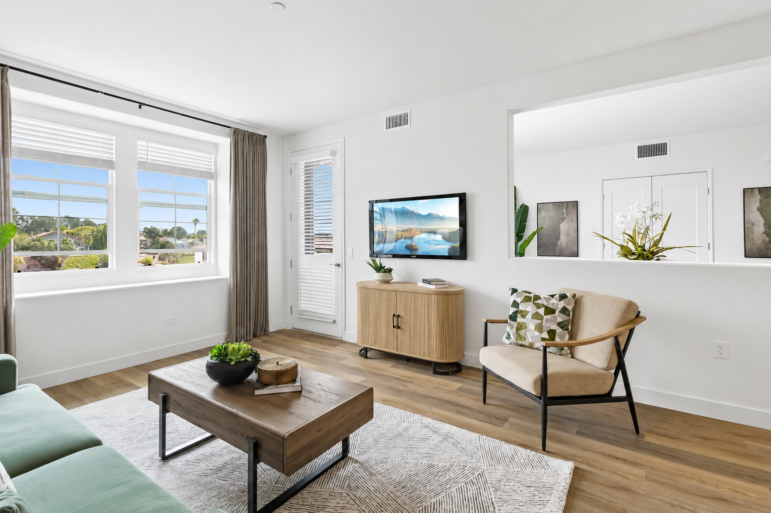 A bright and modern living room featuring a light green sofa, a wooden coffee table with a decorative plant, and a TV mounted on the wall. Large windows allow for natural light, and potted plants add a touch of greenery. The decor includes neutral tones and minimalistic furniture for a cozy atmosphere.